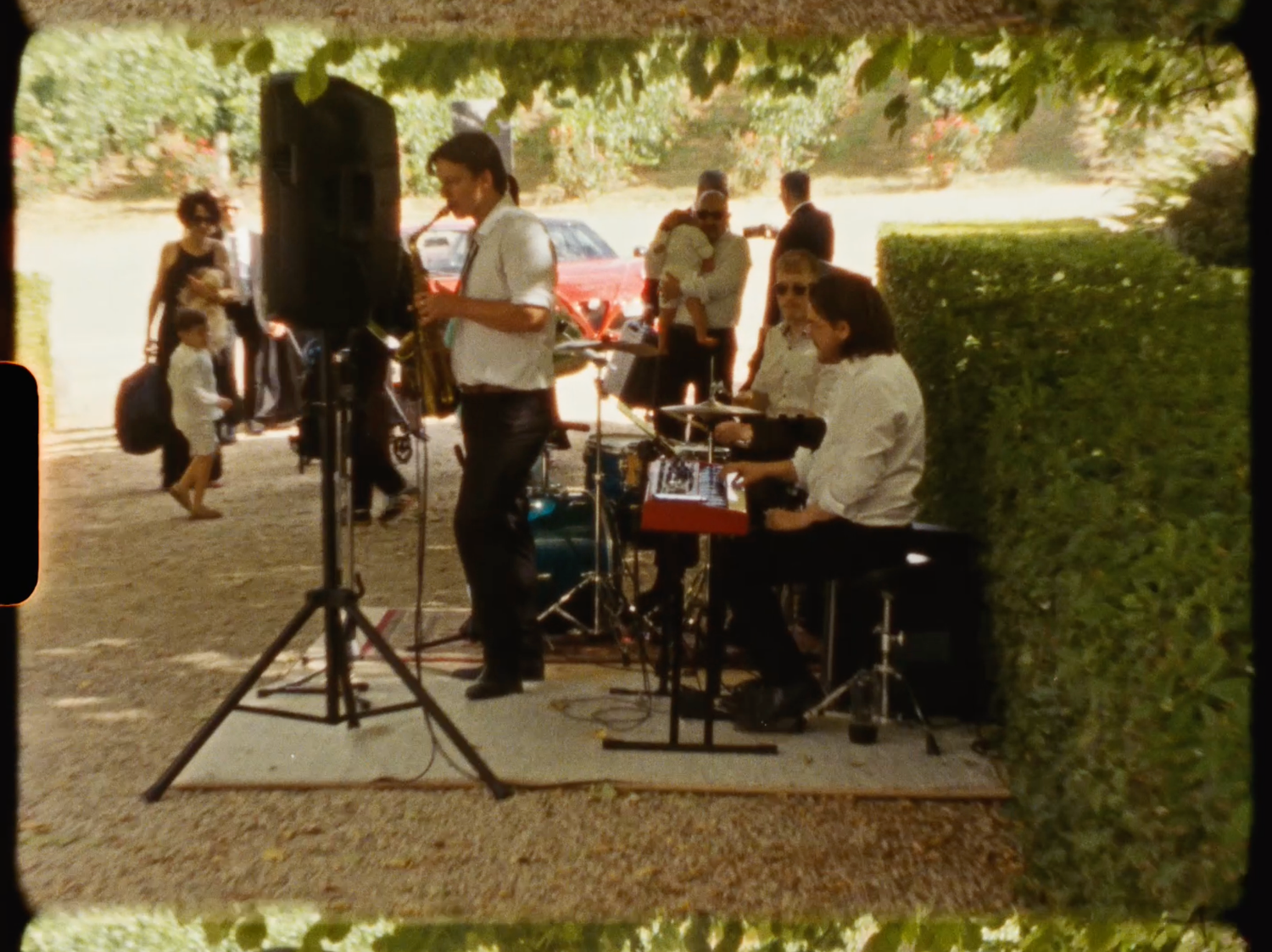 Live band performing outdoors near a hedge at a wedding reception with speakers and drum kit setup.