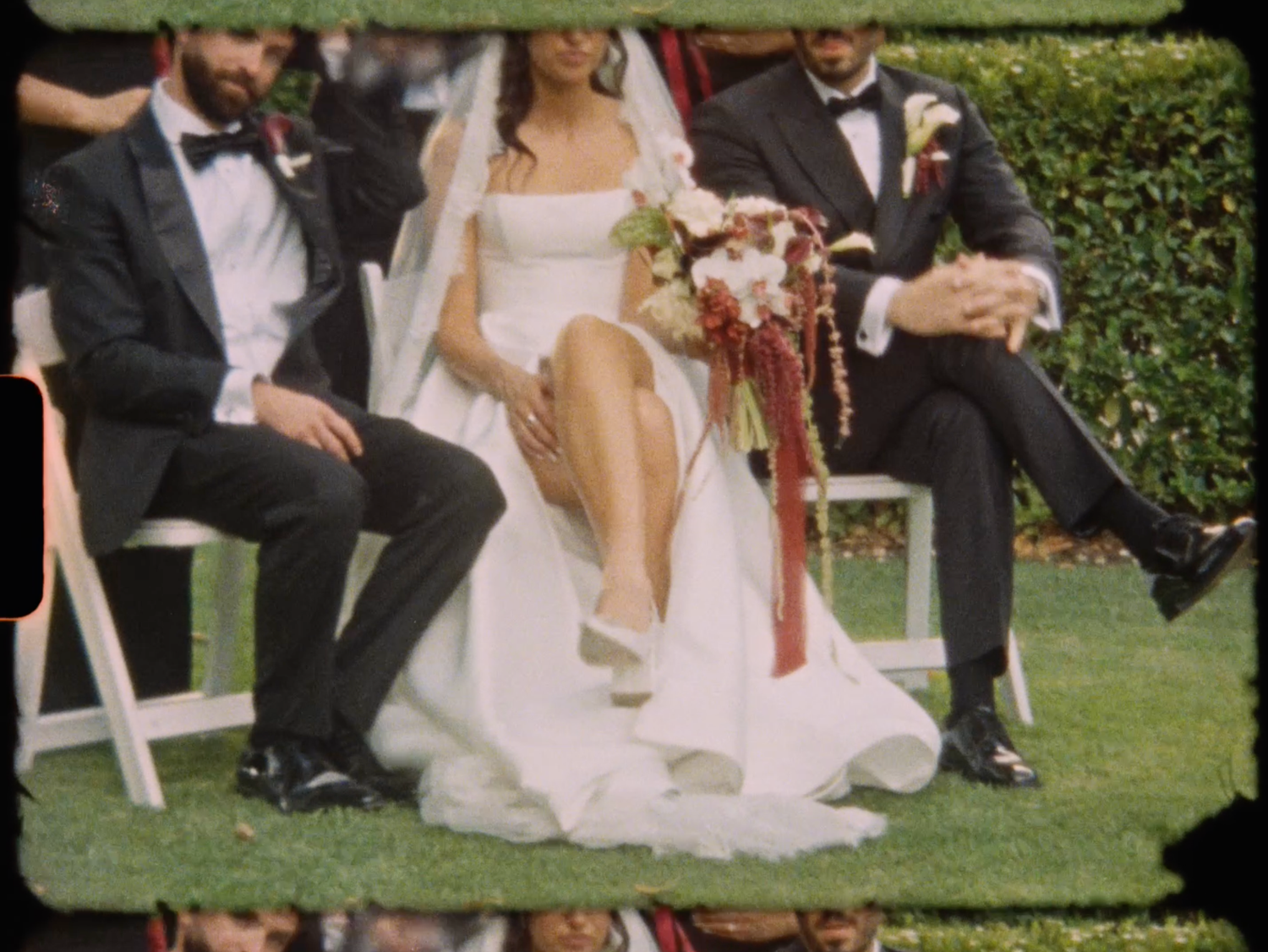 Bride in white gown and groom in tuxedo sit together during outdoor wedding ceremony on green lawn.
