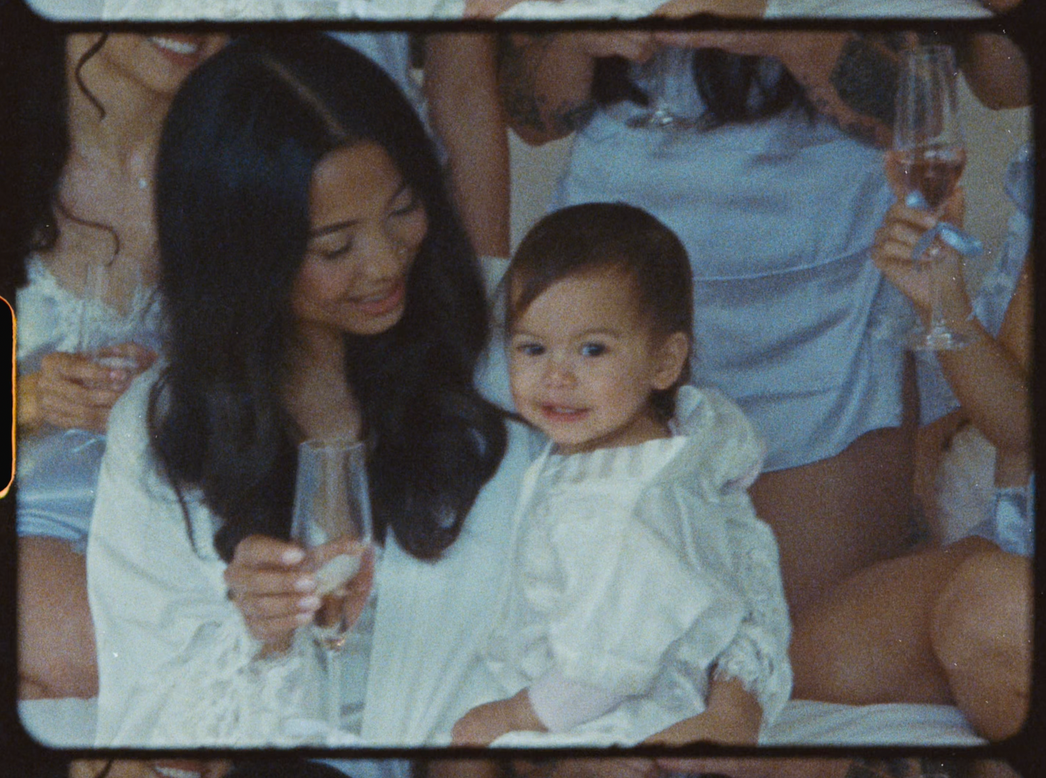 Woman with long dark hair holding champagne glass sitting with smiling toddler in white dress.