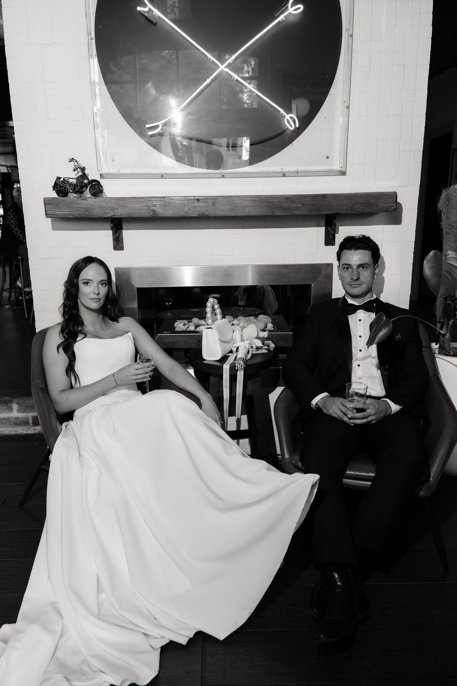 Black and white portrait of bride and groom seated by a fireplace in an elegant venue.