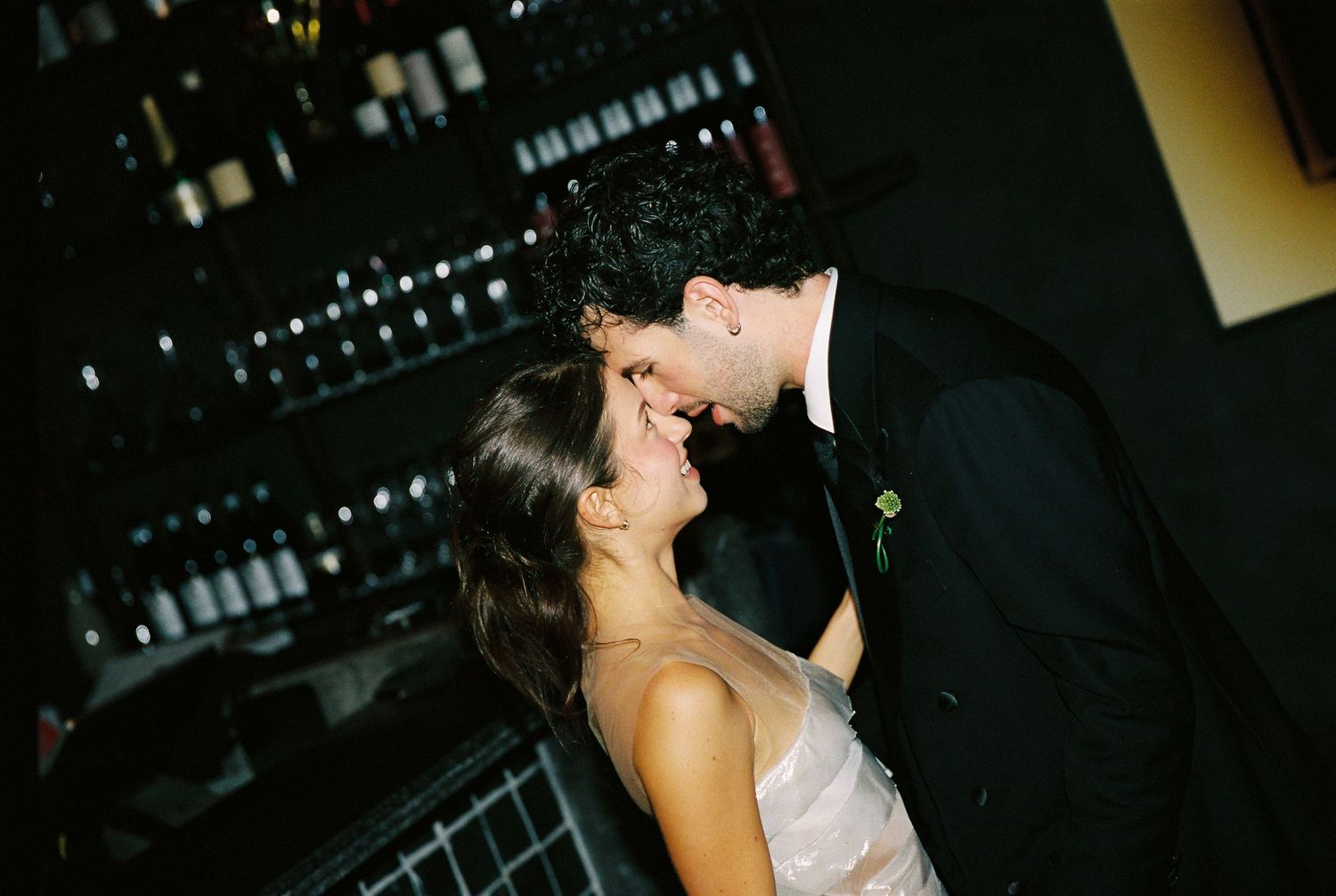 Bride and groom share a romantic almost-kiss moment in dark intimate setting at wedding reception.
