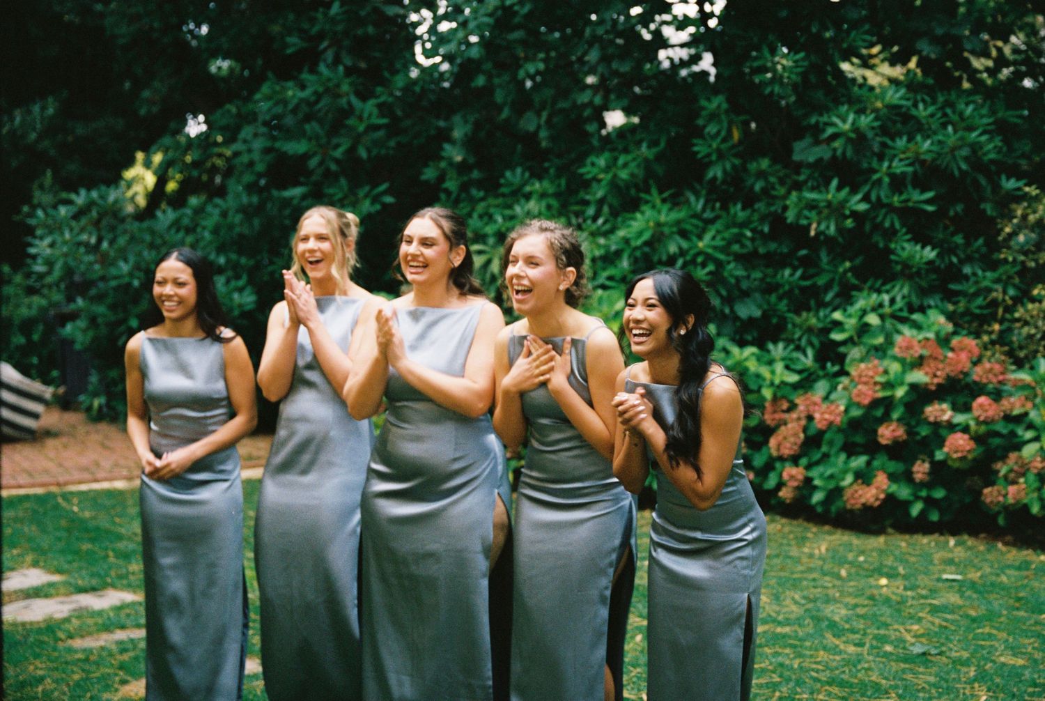 Five bridesmaids in silver satin gowns laughing joyfully outdoors among lush greenery.