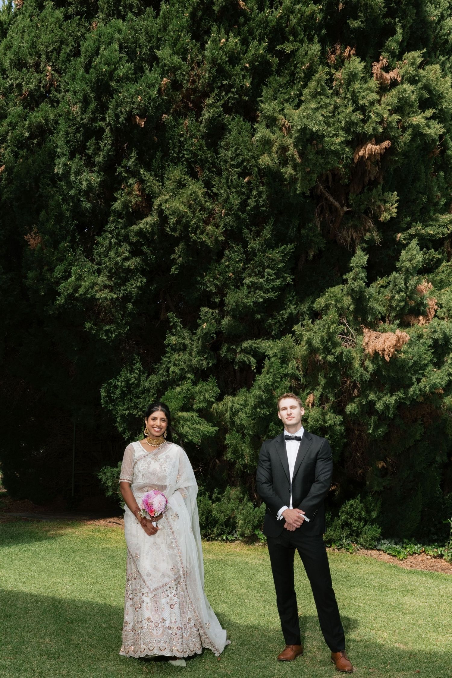 Indian bride in white embroidered lehenga and groom in black tuxedo standing apart in lush garden setting.