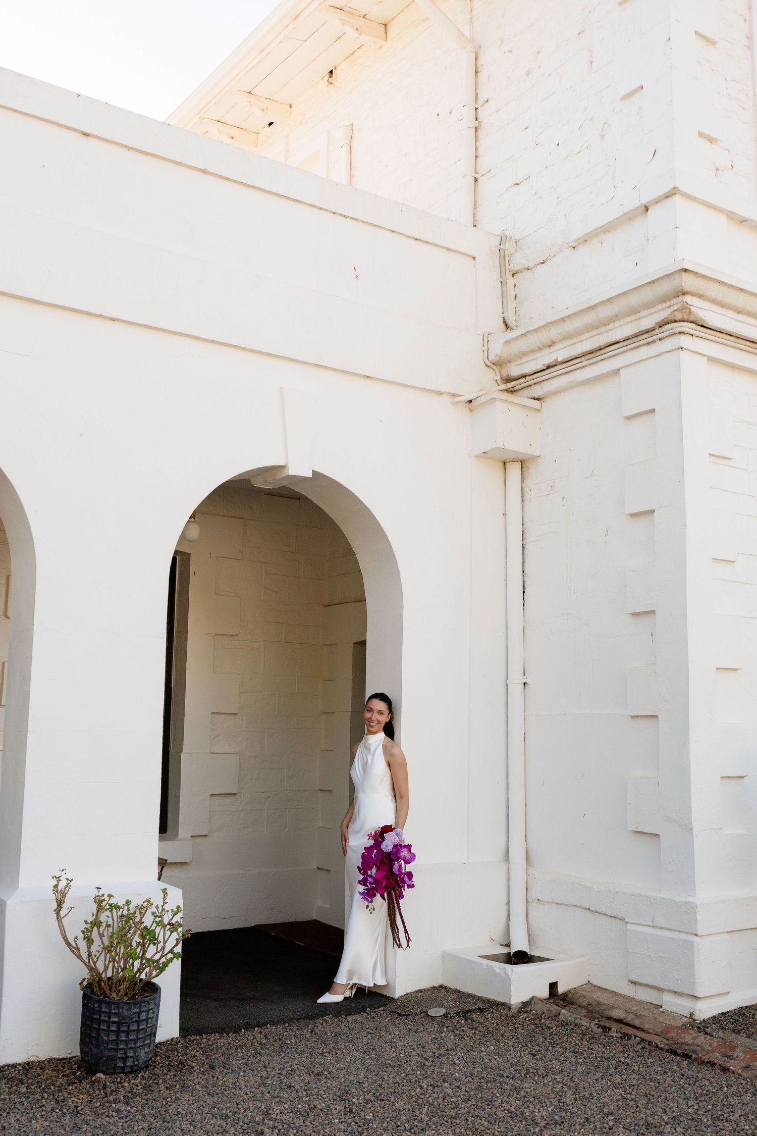 Elegant bride in white halter gown holds purple bouquet beside white arched building.