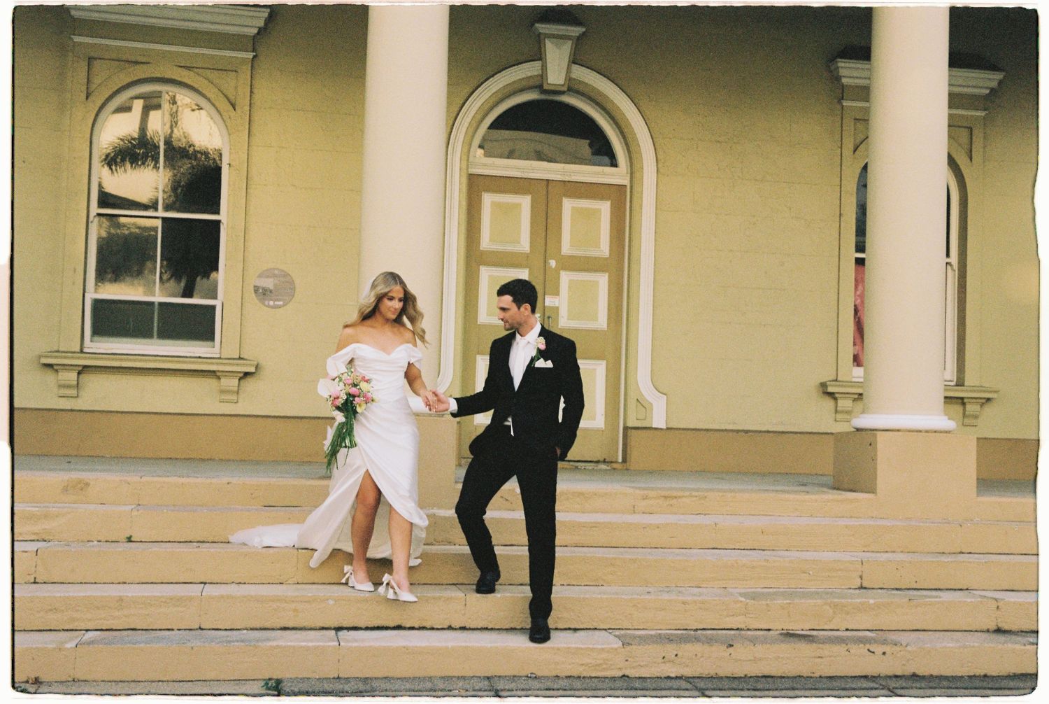 Bride and groom descending steps of a yellow historic building, groom in black tuxedo holding bride's hand.