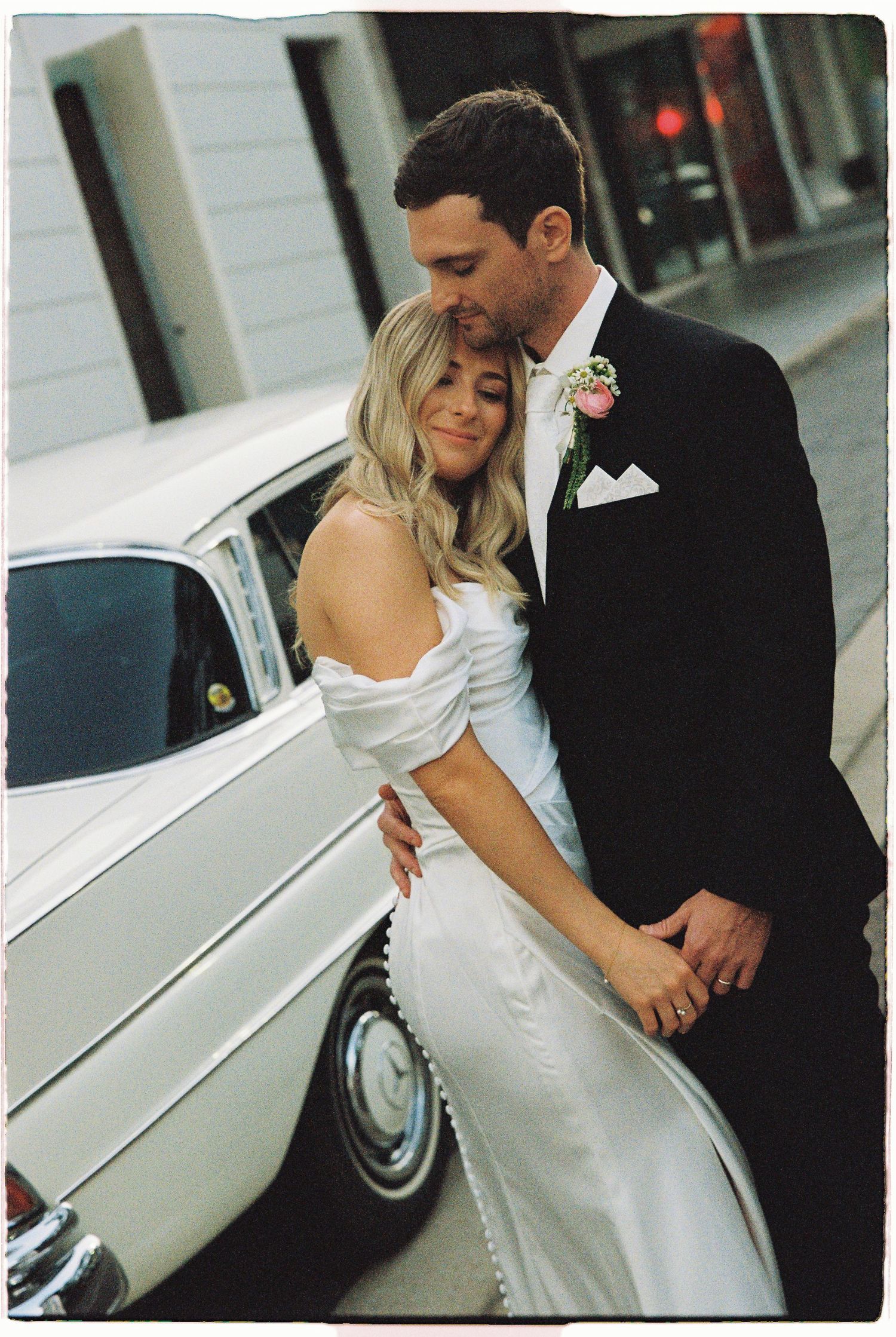 Bride and groom embrace romantically beside a vintage white classic car on a cobblestone street.