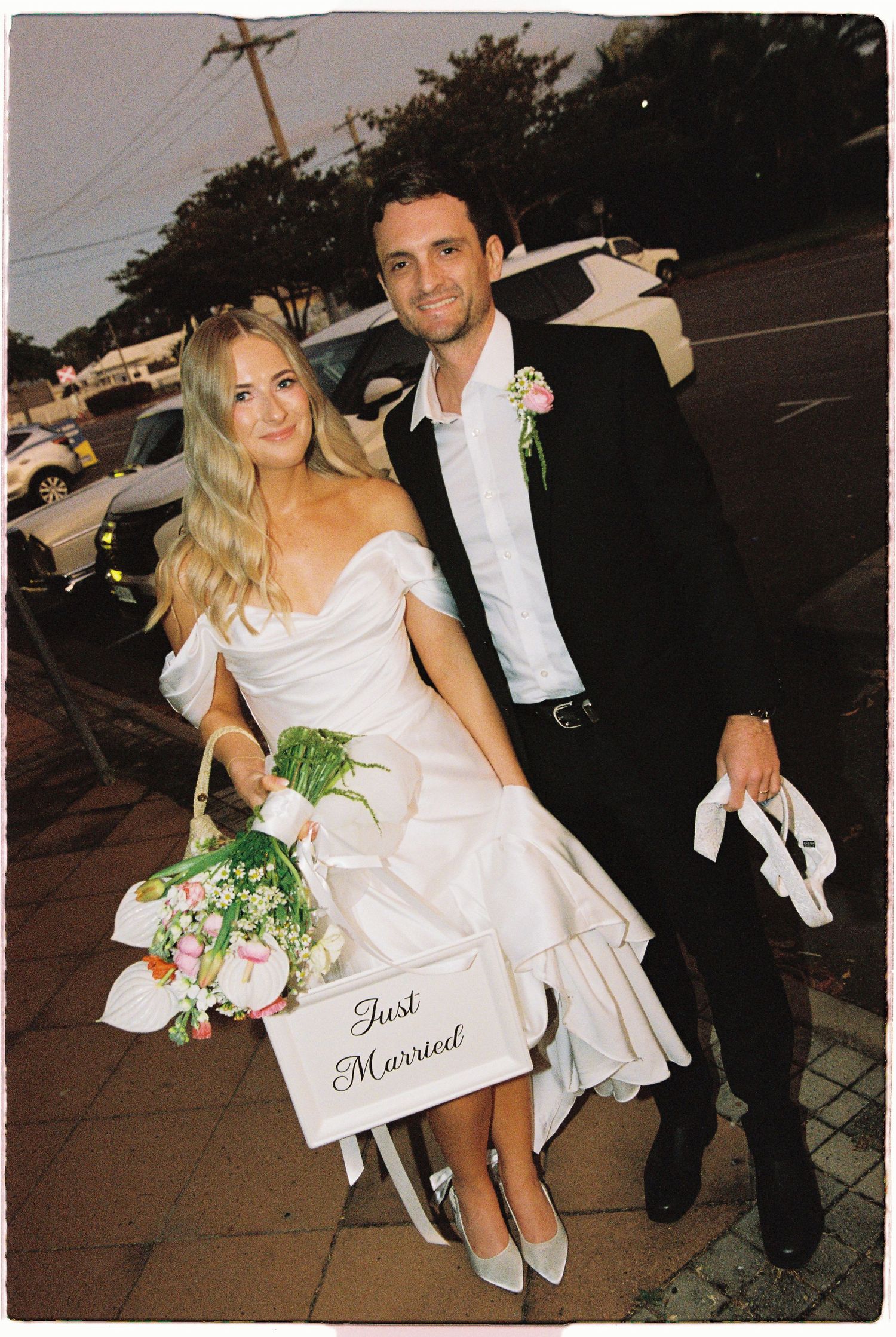 Just married couple holding floral bouquet and sign on sidewalk, bride in white gown, groom in black suit.