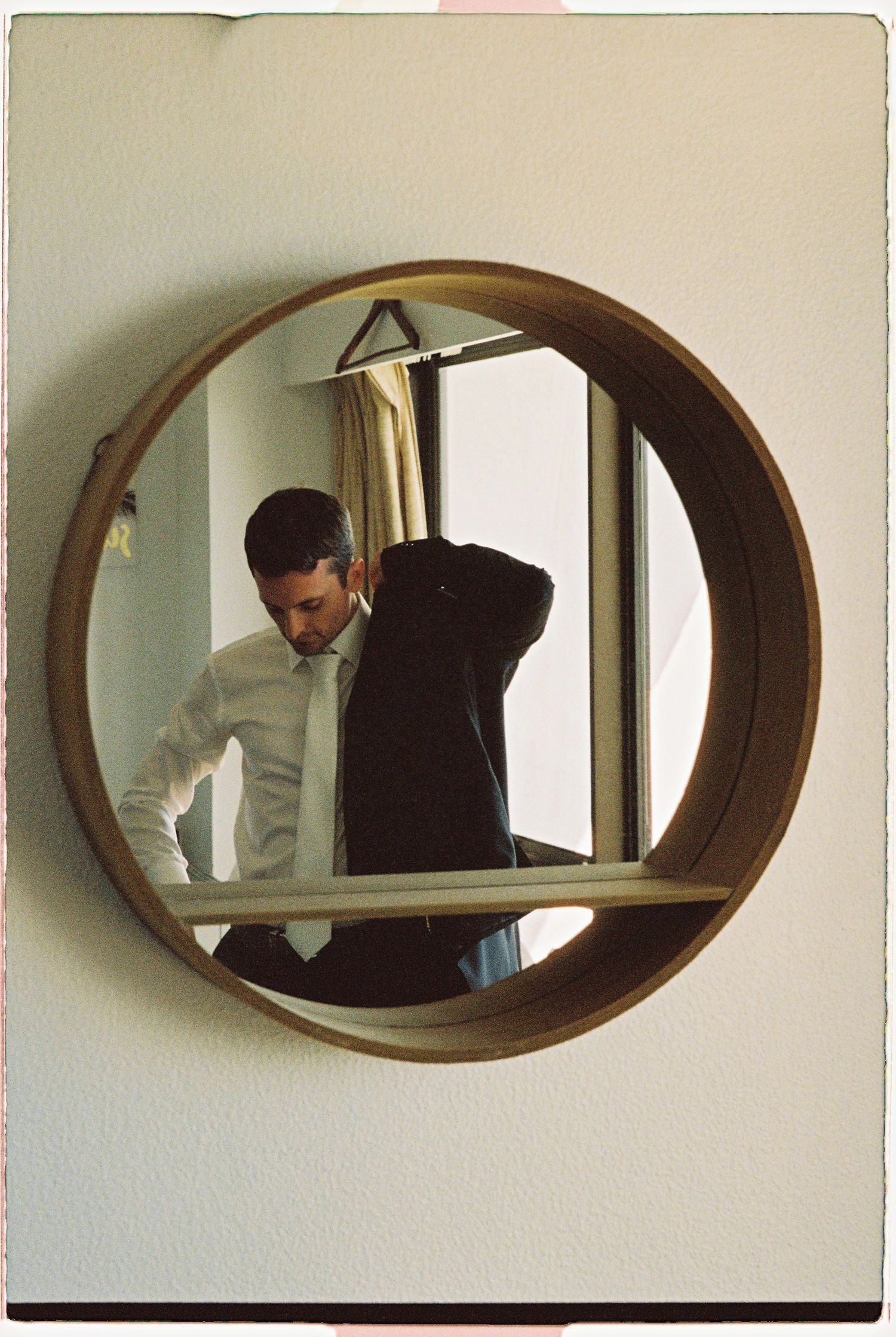 Groom adjusting his suit jacket reflected in a round circular wall mirror in a hotel room.