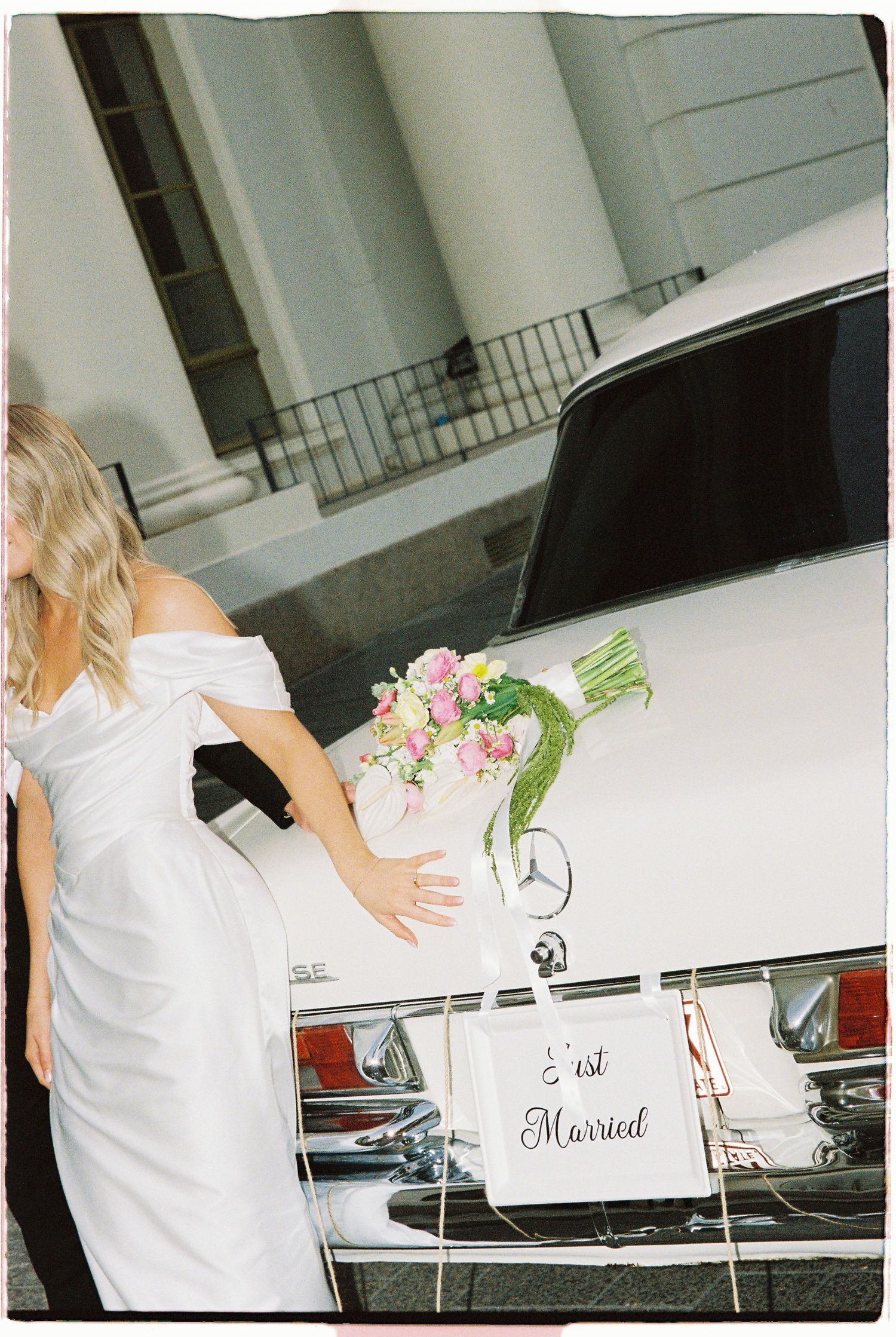 Bride in white off-shoulder gown holding pink bouquet beside vintage white Mercedes with Just Married sign.