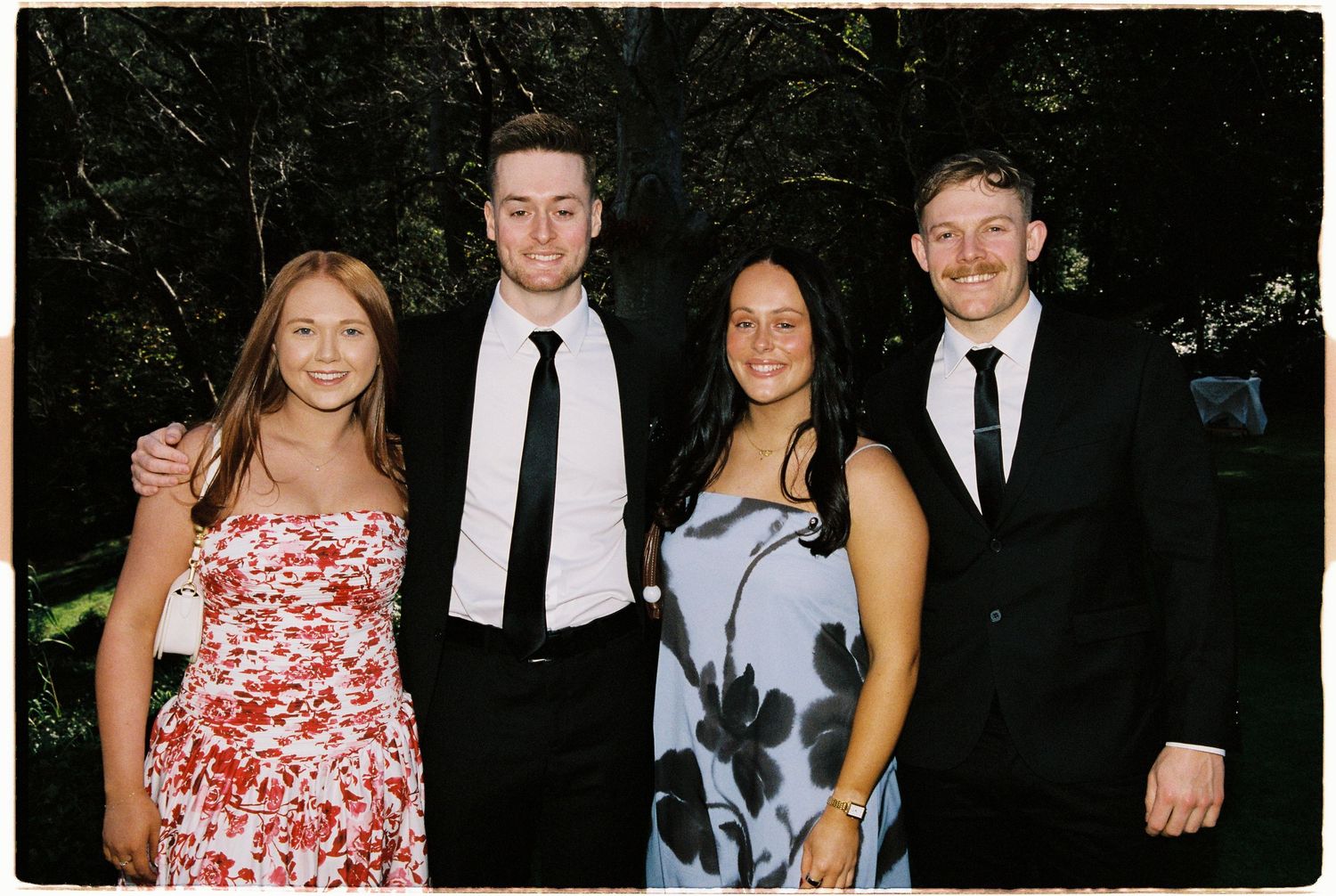 Two couples dressed in formal attire smiling together outdoors at a wedding celebration.