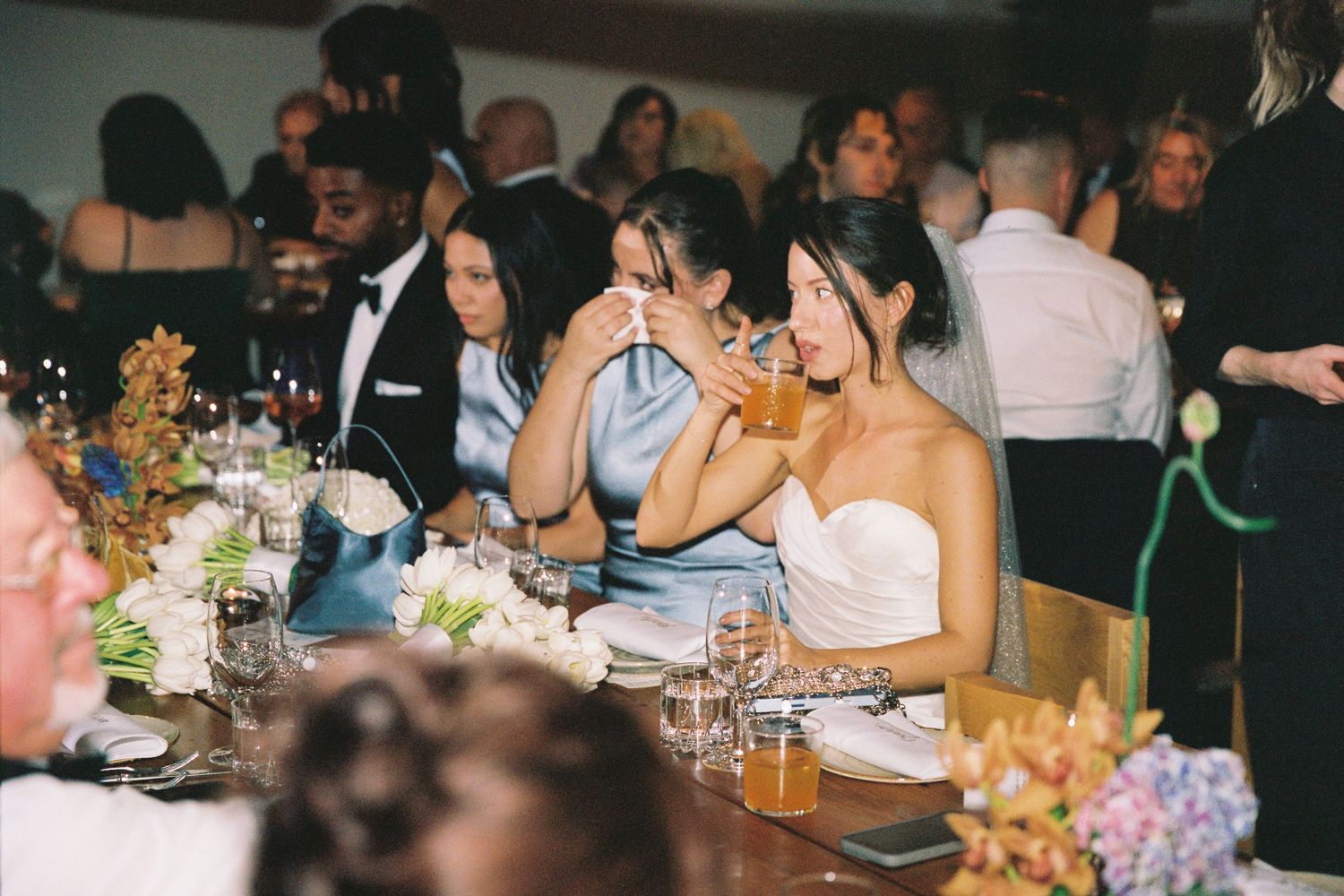 Elegantly dressed wedding guests sip cocktails at a candlelit reception dinner table adorned with floral centerpieces.