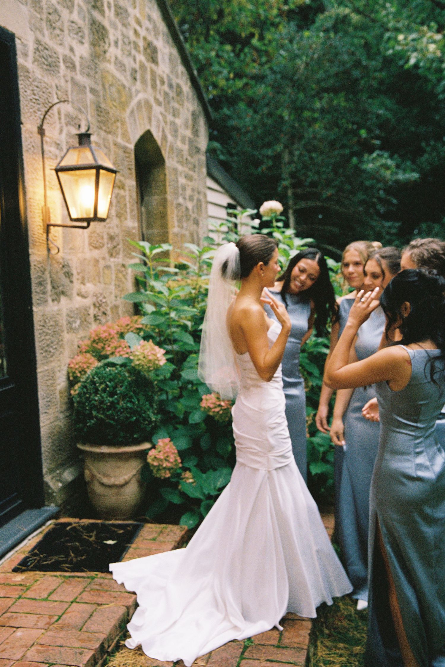 Bride in white mermaid gown with veil shares joyful moment with bridesmaids in blue dresses outside stone building.