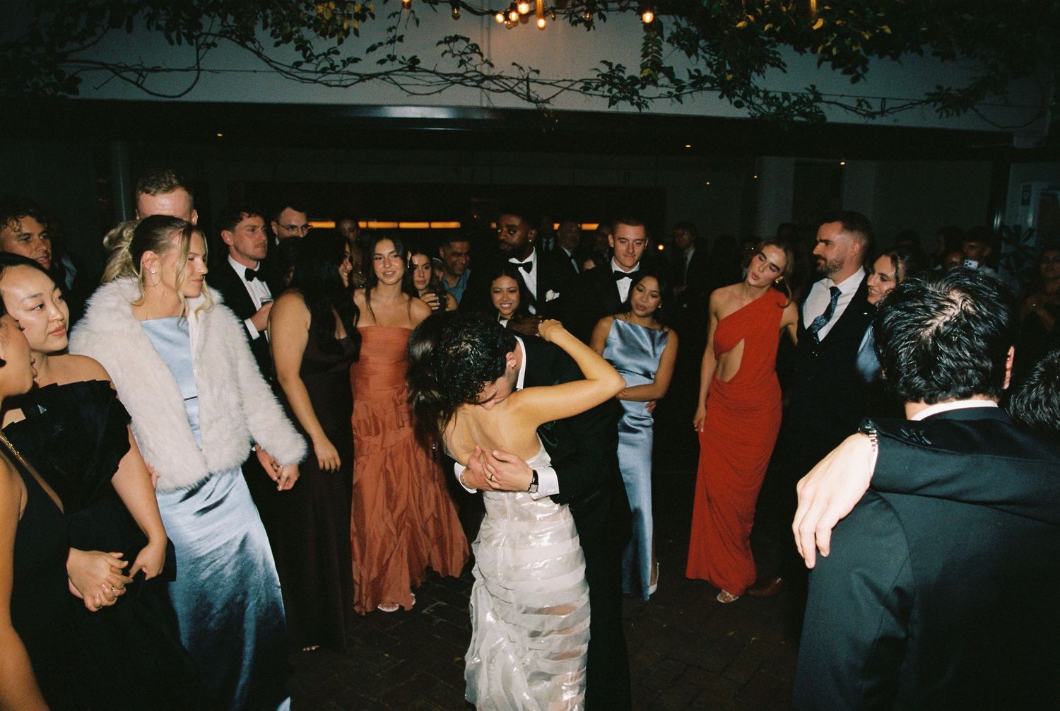 Bride and groom share first dance surrounded by elegantly dressed wedding guests on a lively reception dance floor.