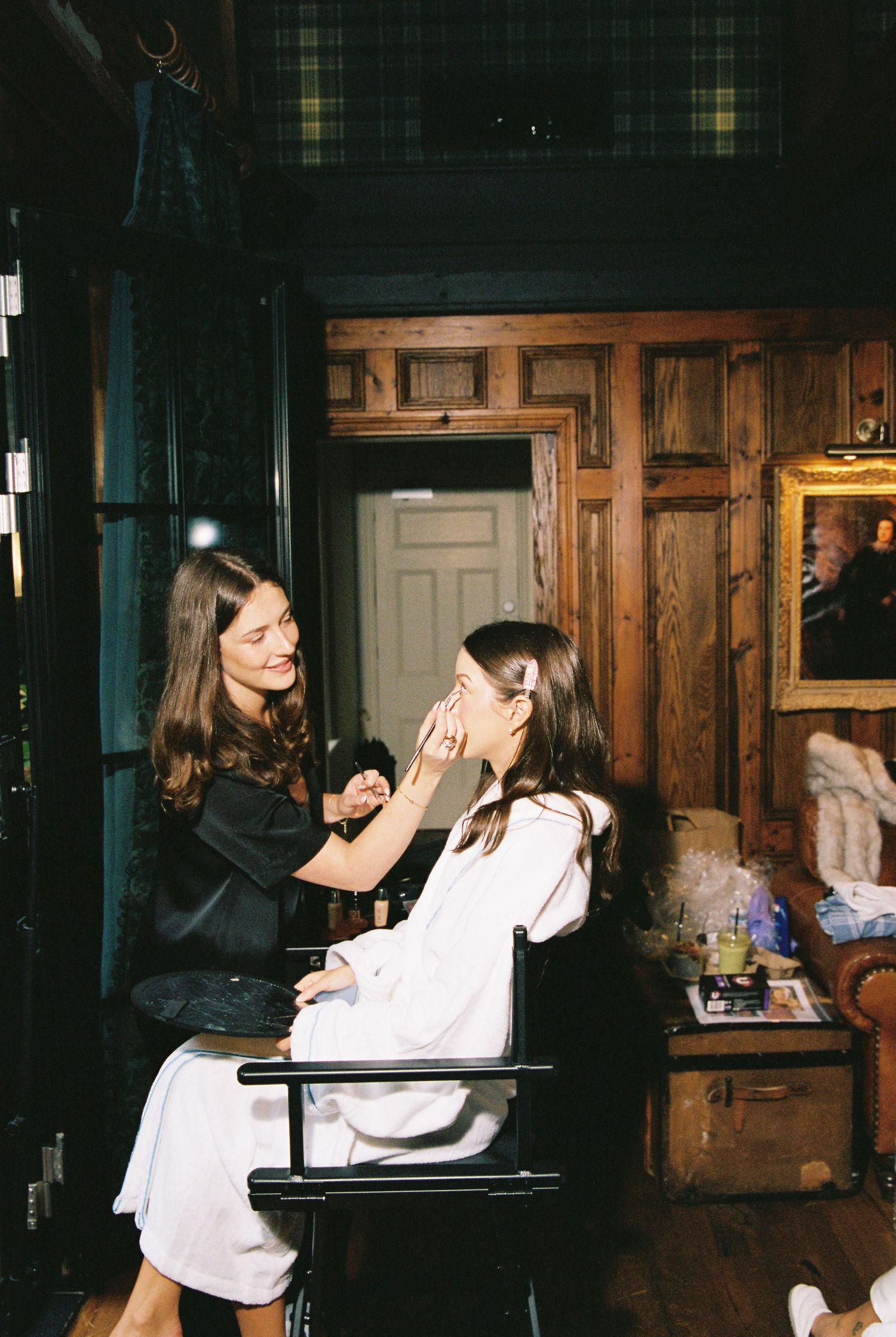 Makeup artist in black applying bridal makeup to a woman in white robe in a dark wood-paneled room.