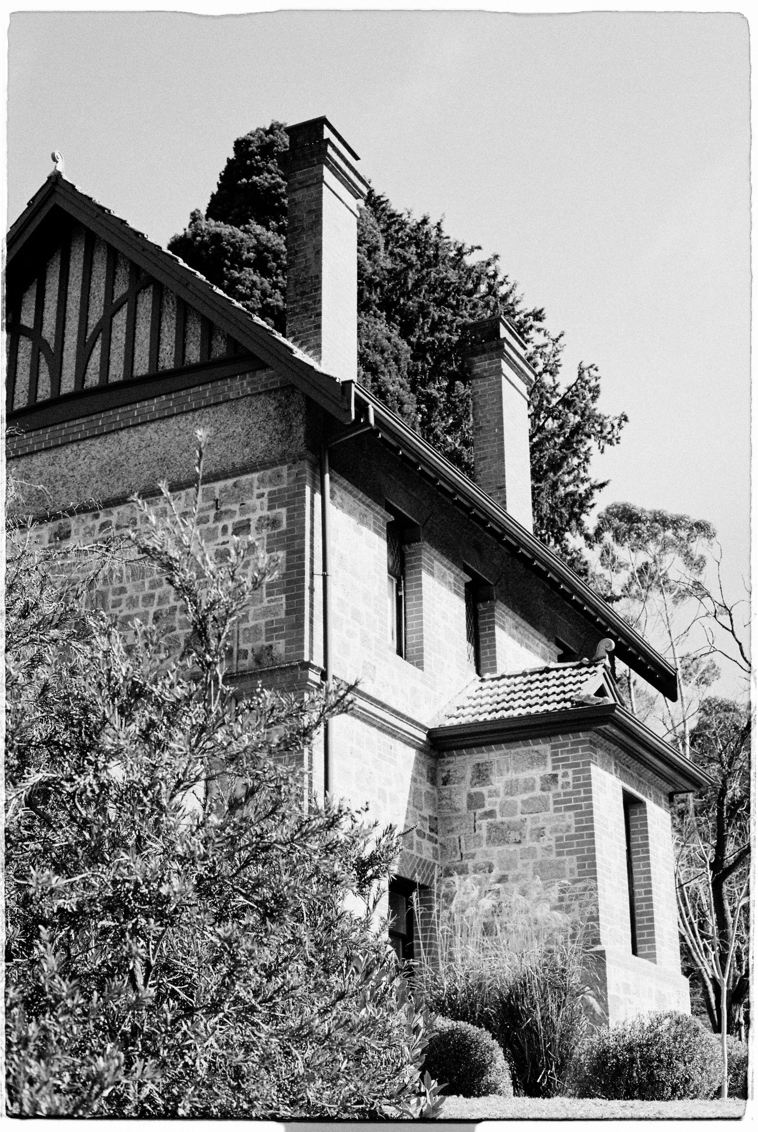 Black and white photo of a historic stone mansion with Tudor-style gabled roof, chimney, and lush garden surroundings.