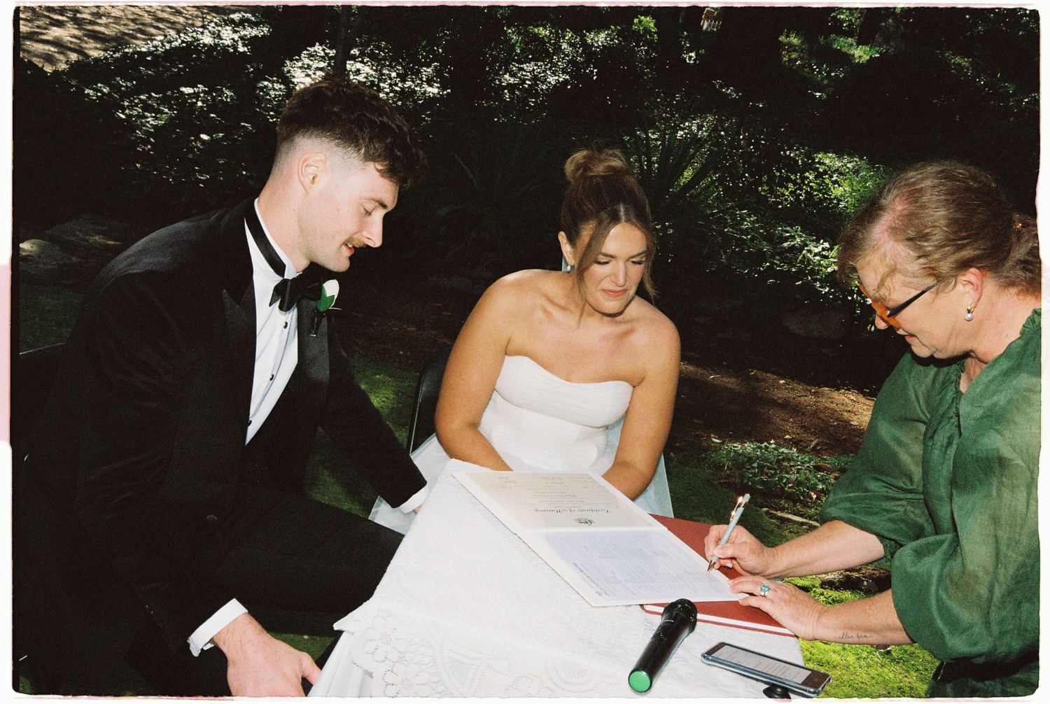 Bride and groom signing wedding documents with officiant outdoors, surrounded by lush green trees.