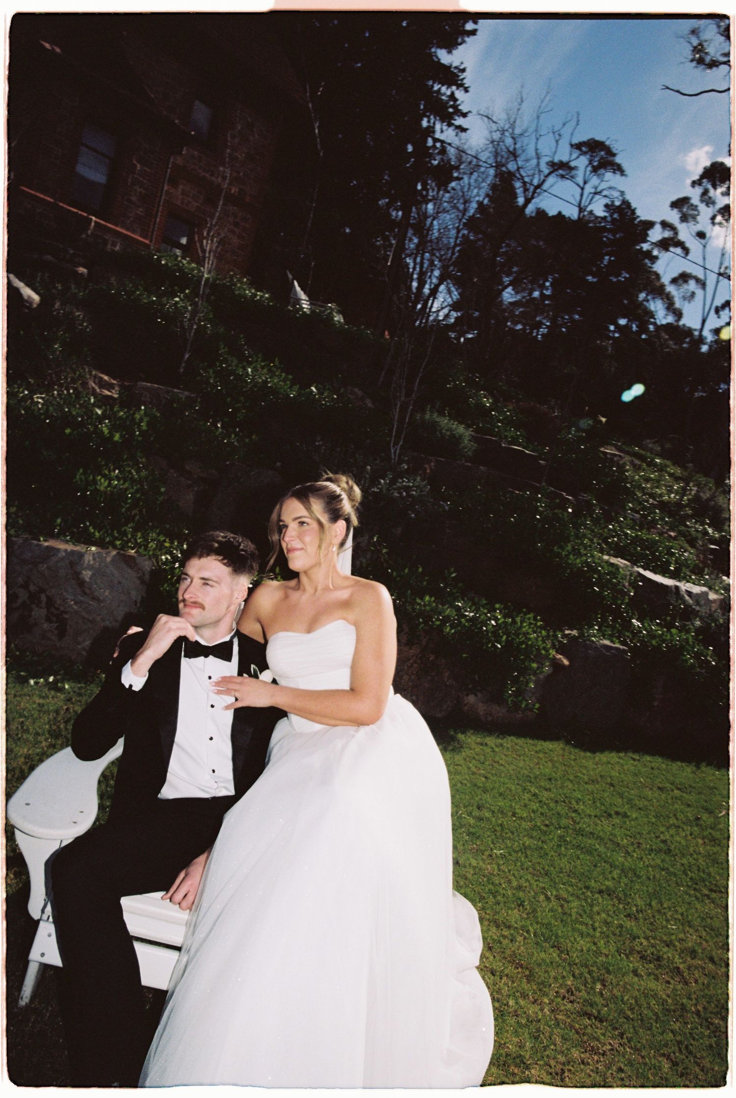 Bride and groom sitting on white outdoor chair at wedding venue with lush green hillside backdrop.