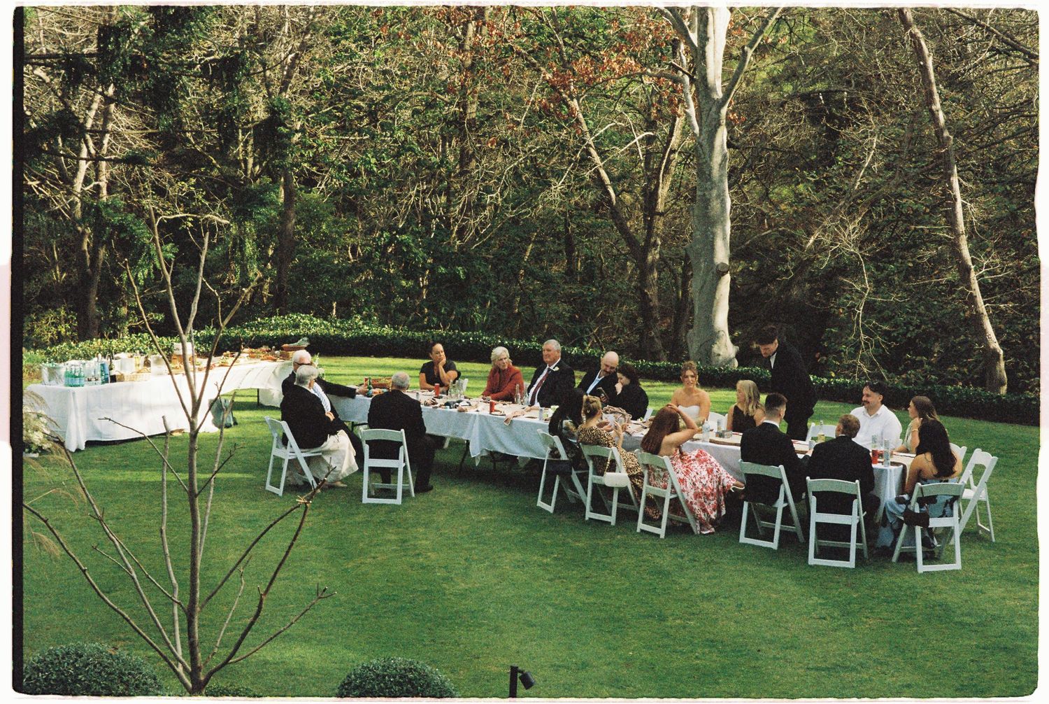 Elegant outdoor wedding reception with guests seated at long white tables on green lawn.