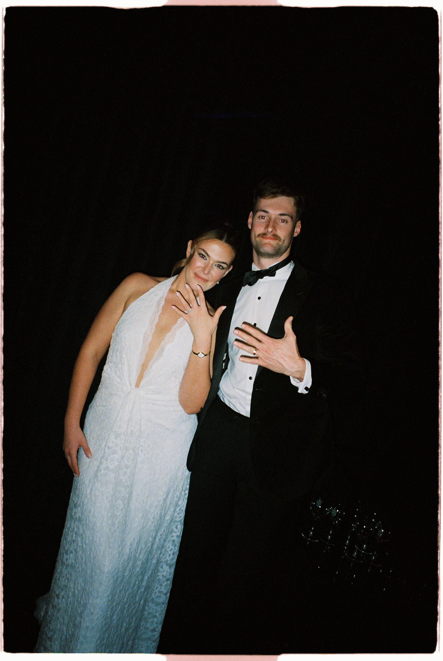 Bride and groom posing against dark background, bride showing wedding ring in white gown.