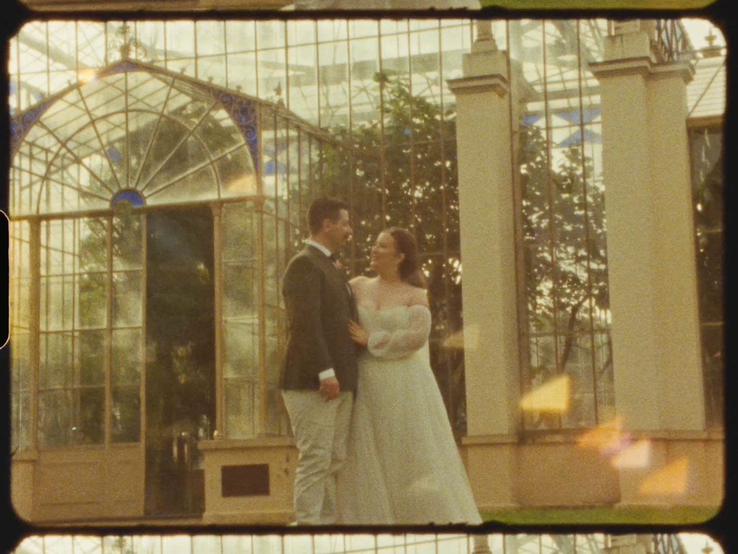 A bride and groom stand together smiling outside a glass greenhouse venue.