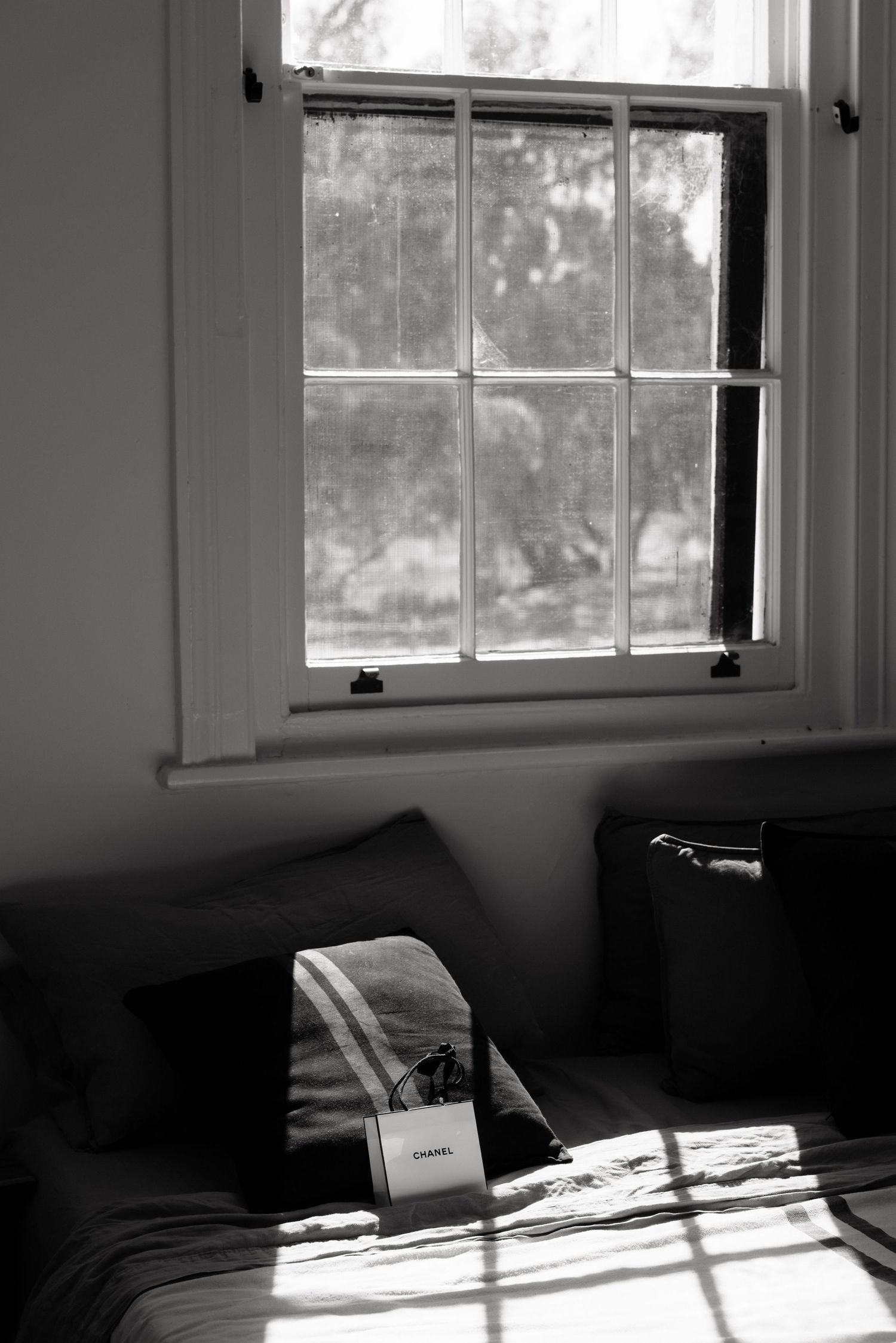 Sunlight streams through a vintage multi-pane window onto a bed with a Chanel bag in black and white.