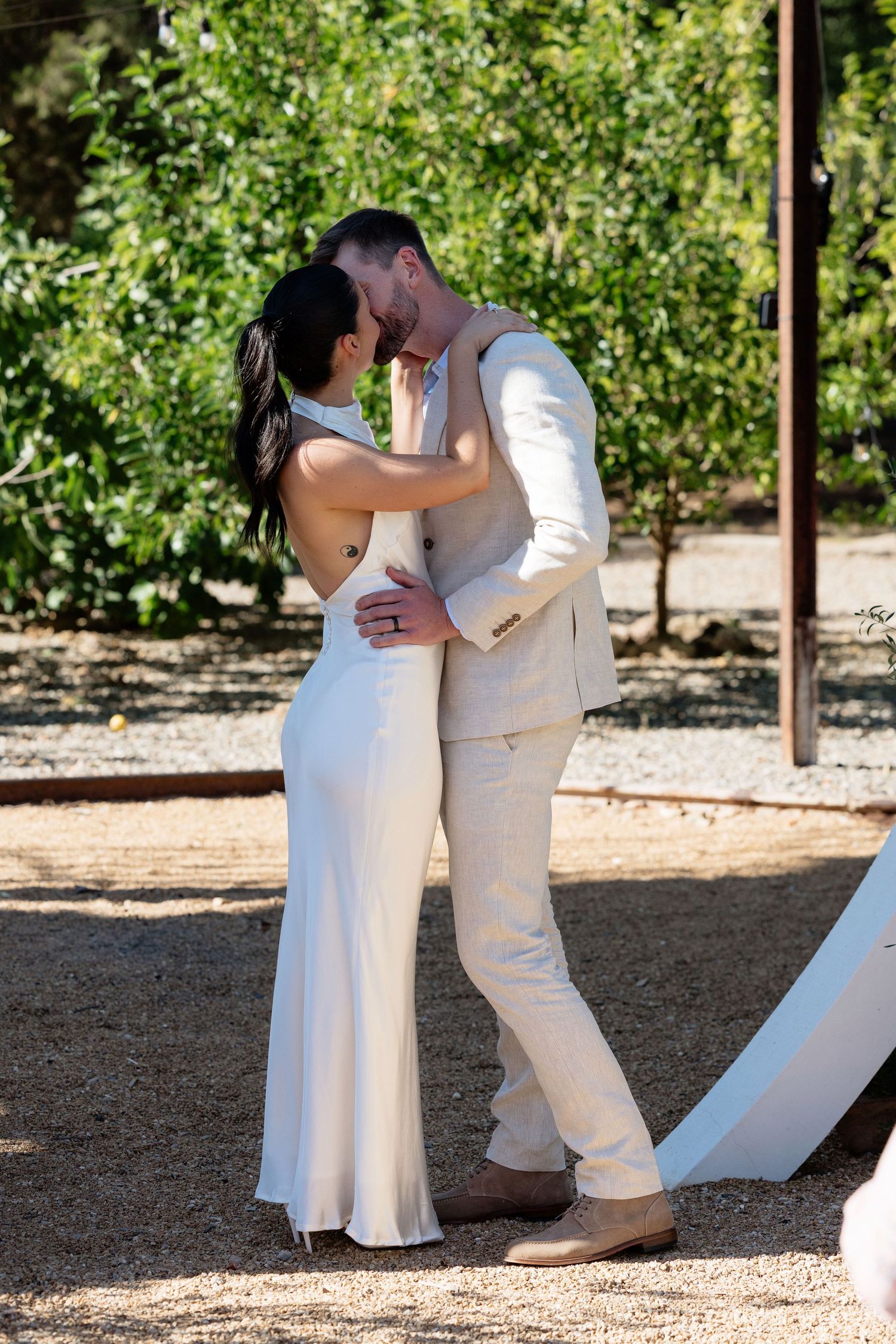 Bride and groom share first kiss in outdoor garden setting, bride in white gown, groom in beige suit.