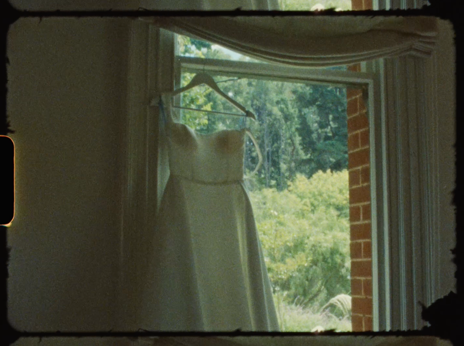 Vintage film-style photo of a white wedding dress hanging in a sunlit bay window.