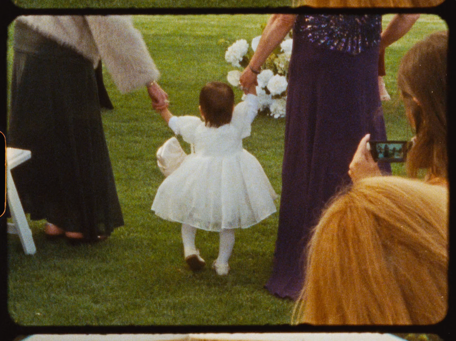 Flower girl in white dress walking down the aisle at an outdoor garden wedding ceremony.