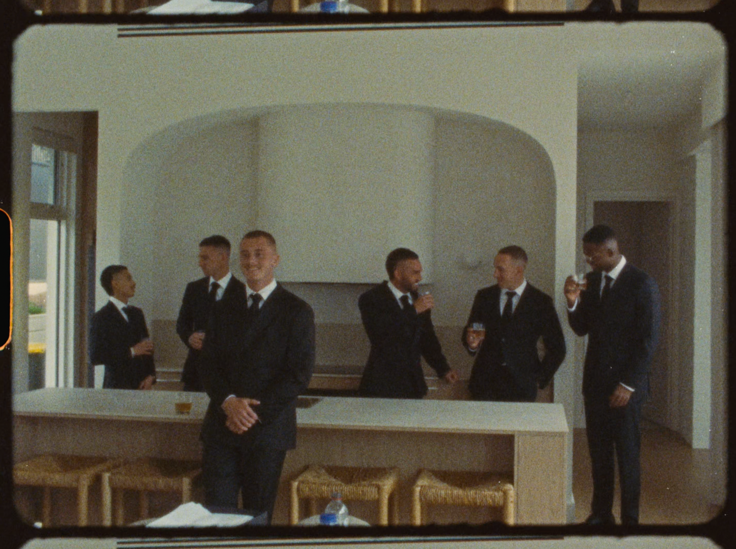 Group of groomsmen in black suits gathered in modern kitchen space before wedding ceremony.