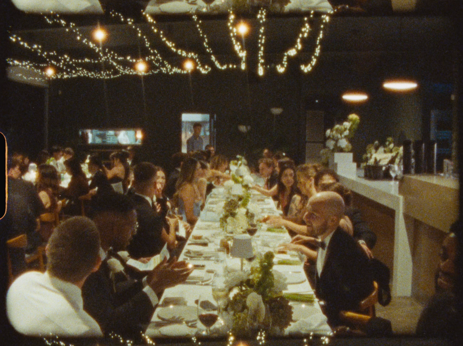 Guests gather at a long decorated dinner table under fairy lights at a wedding reception.
