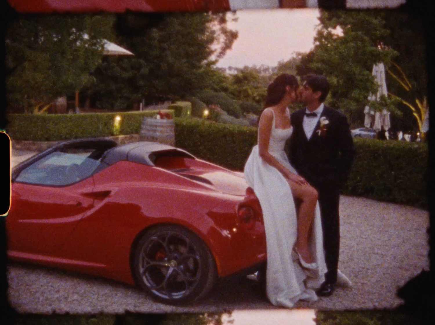 Bride and groom sharing a kiss beside a red Ferrari sports car during outdoor wedding celebration.