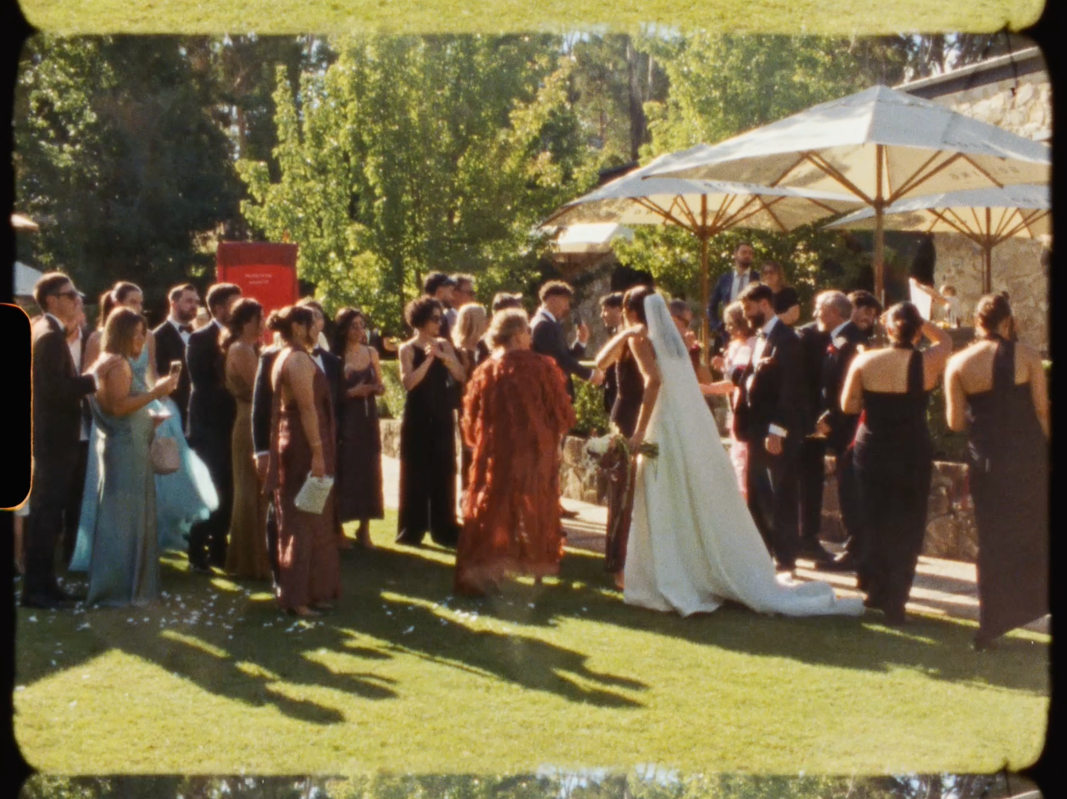 Wedding guests mingle on a sunlit lawn as the bride in a flowing white gown greets attendees.