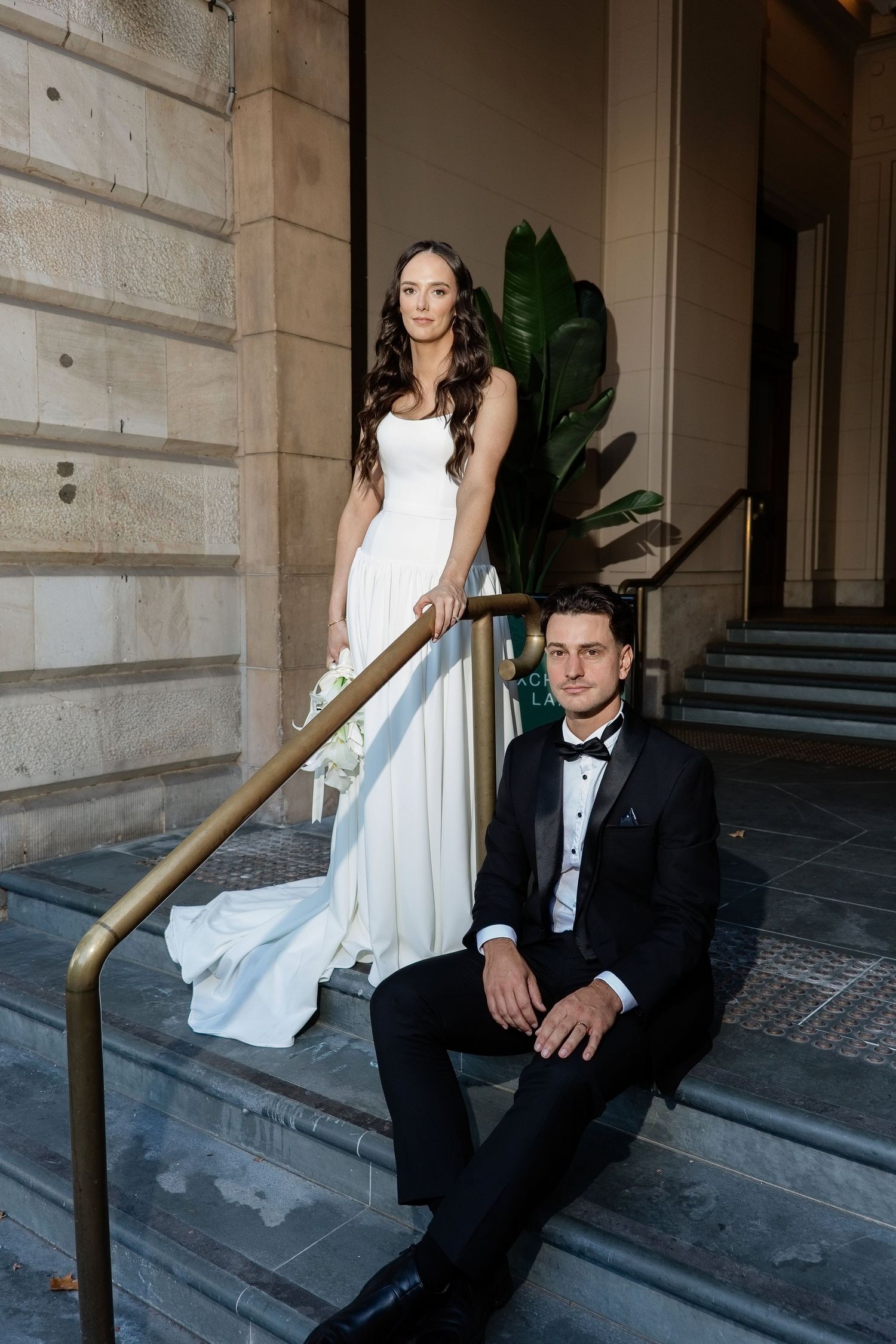 Bride standing on steps holding bouquet with groom seated beside a gold railing.