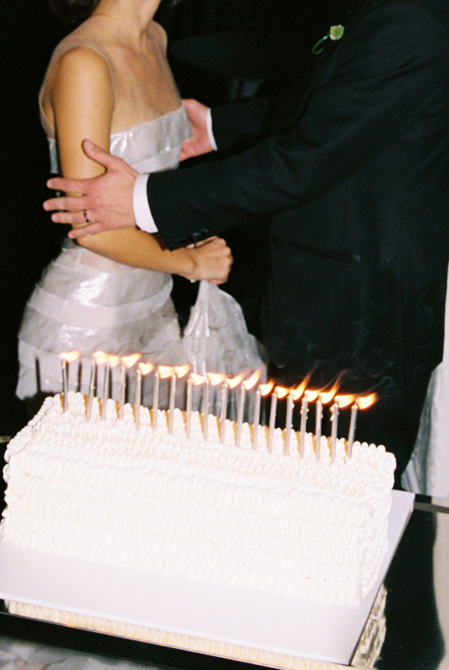 Bride and groom cutting white wedding cake adorned with lit candles at their reception.