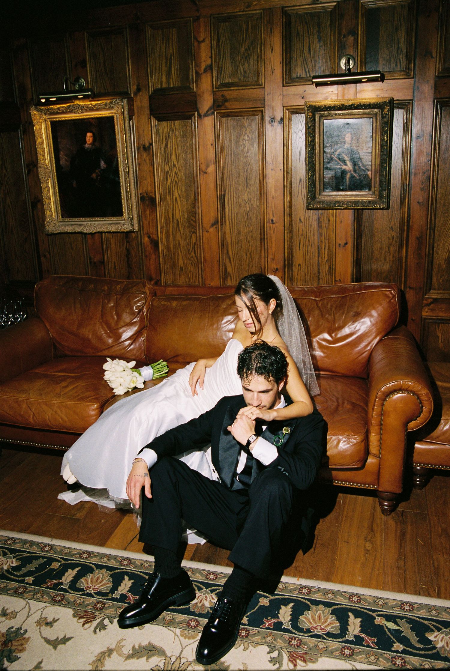 Groom in black tuxedo sits with bride in white gown and veil on leather sofa in wood-paneled room.