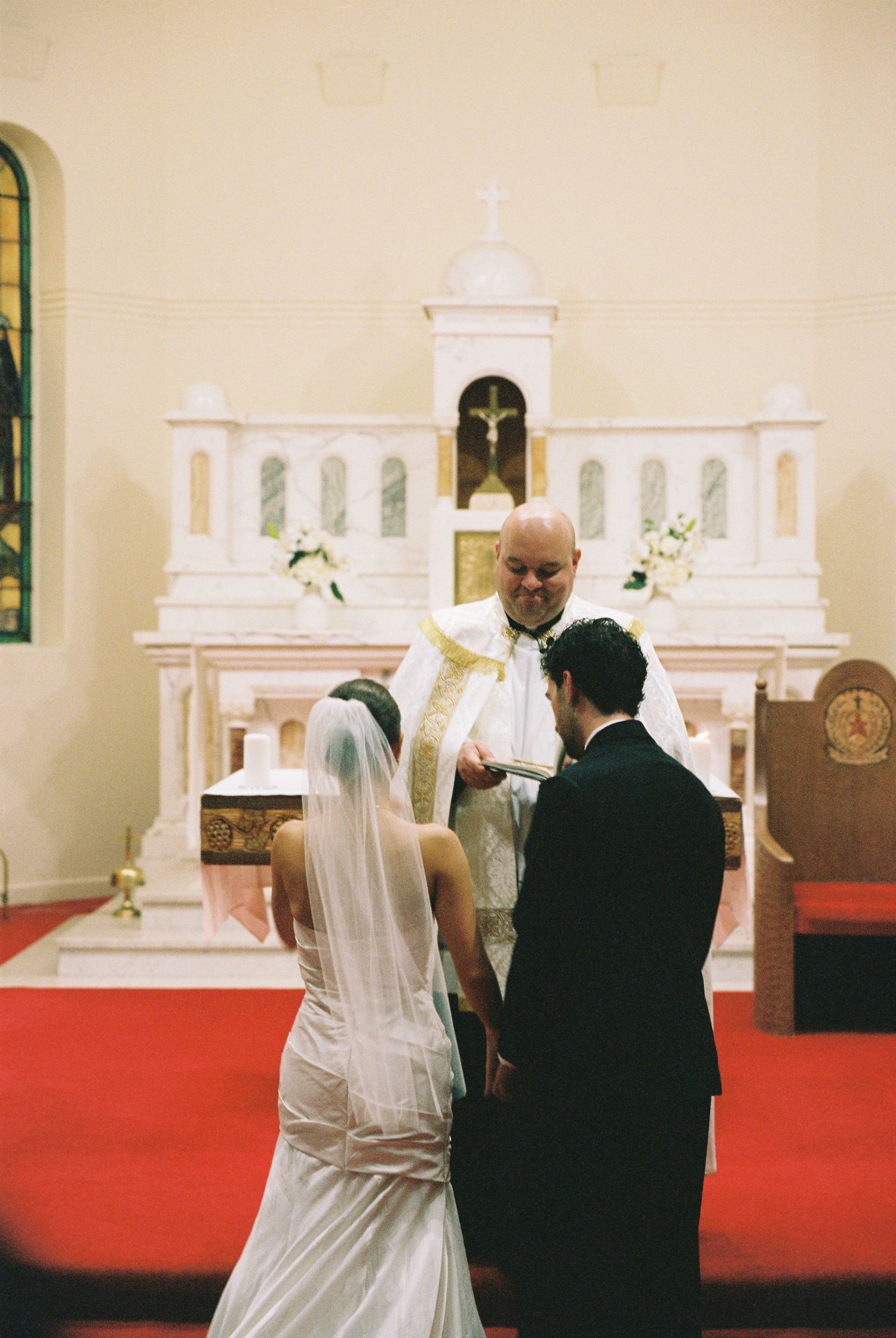 Bride and groom exchange vows before a priest at a white Catholic church altar.