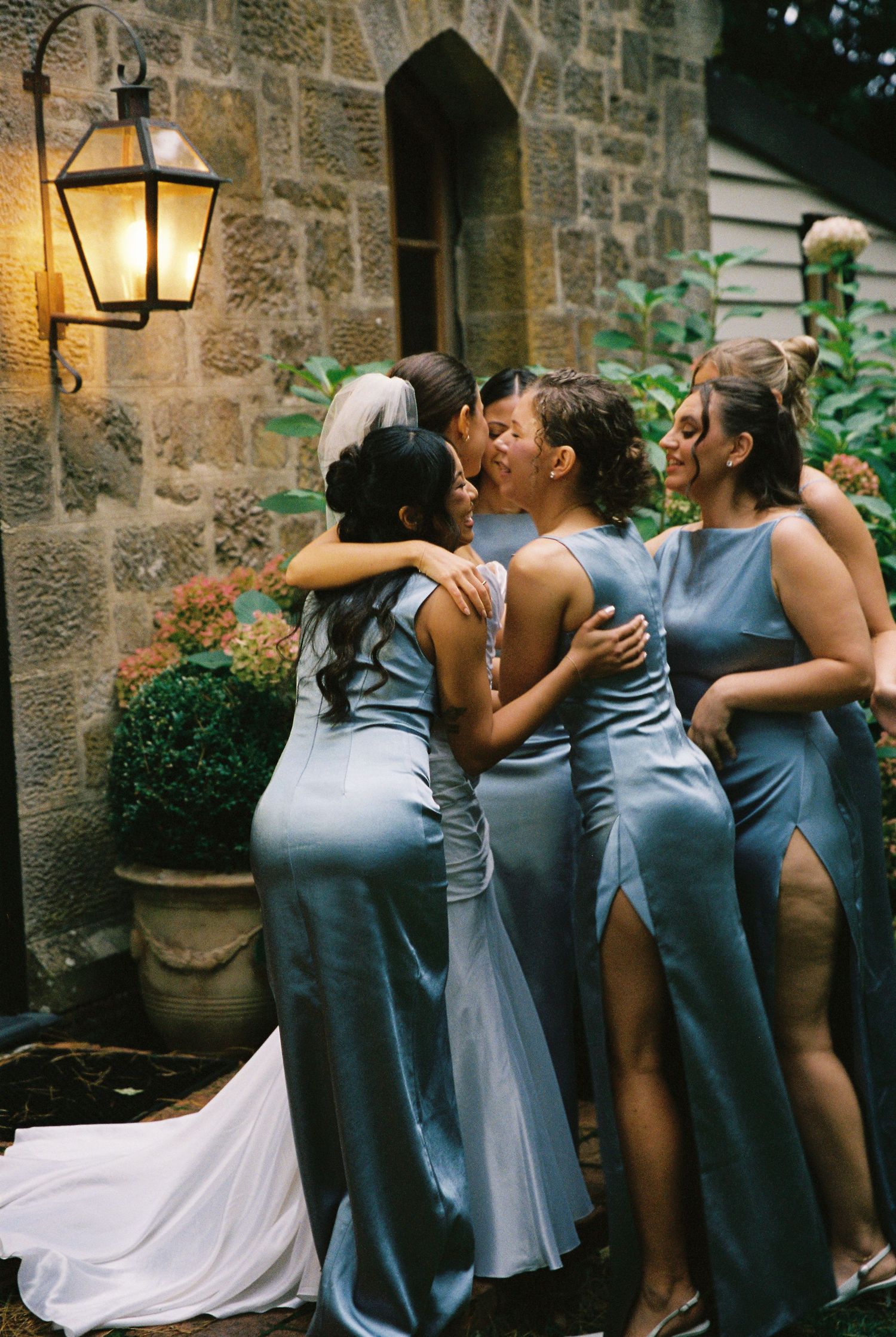 Bride in white gown embracing bridesmaids in dusty blue satin dresses outside stone building at night.