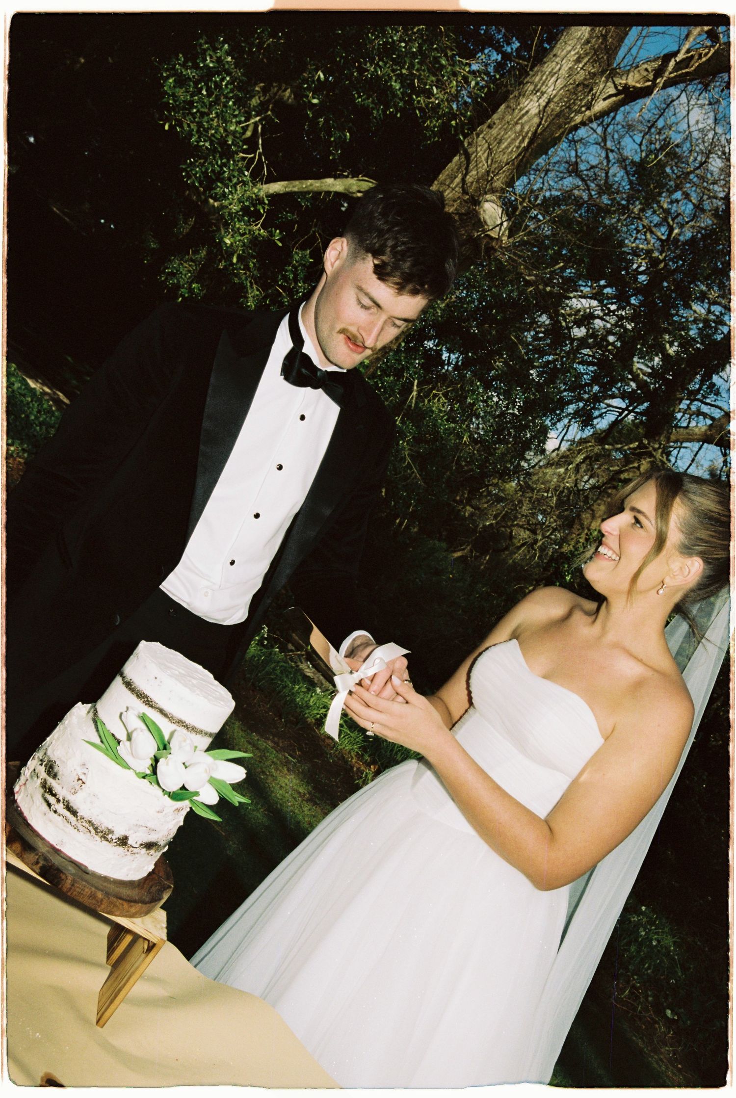 Bride and groom cut rustic naked wedding cake outdoors under trees, groom in black tuxedo.