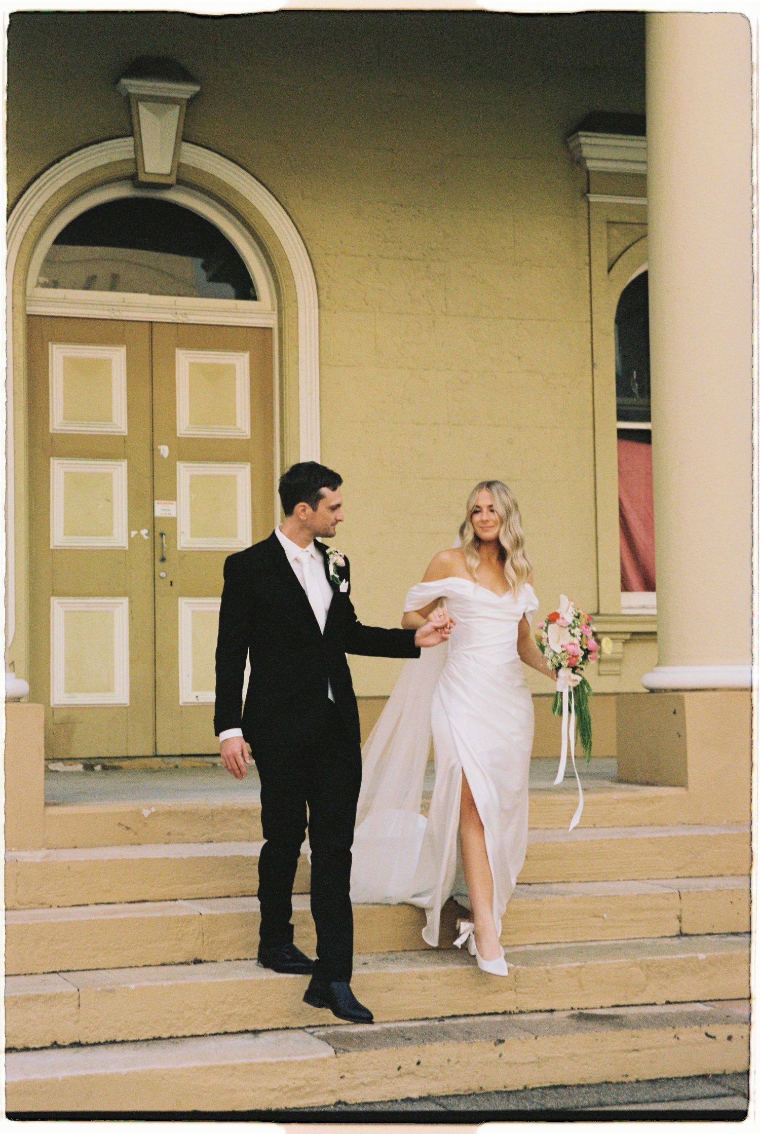 Bride and groom walk down church steps, she holds a colorful bouquet in an off-shoulder white gown, he wears a black tuxedo.