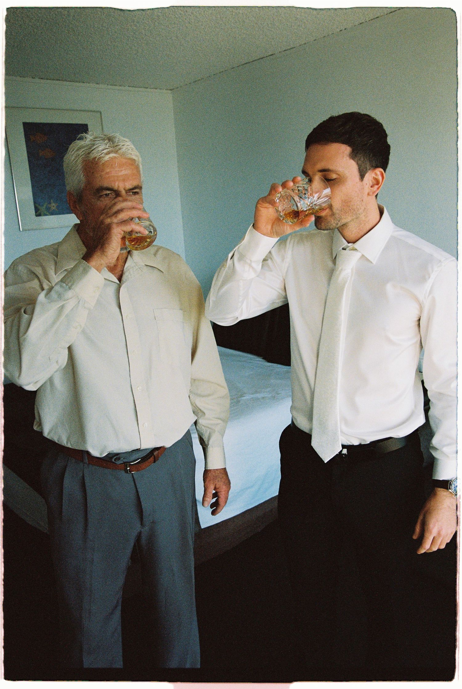 Two men in white dress shirts and ties drinking whiskey together in a hotel room.