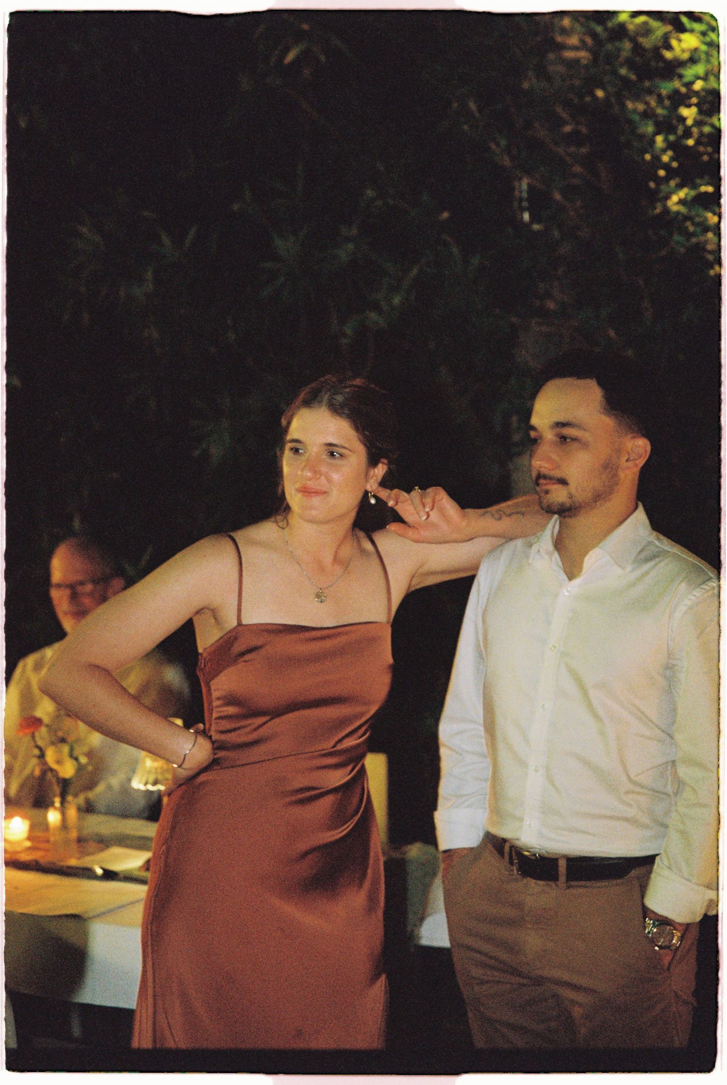 Woman in rust satin slip dress and man in white shirt posing at outdoor evening wedding reception.