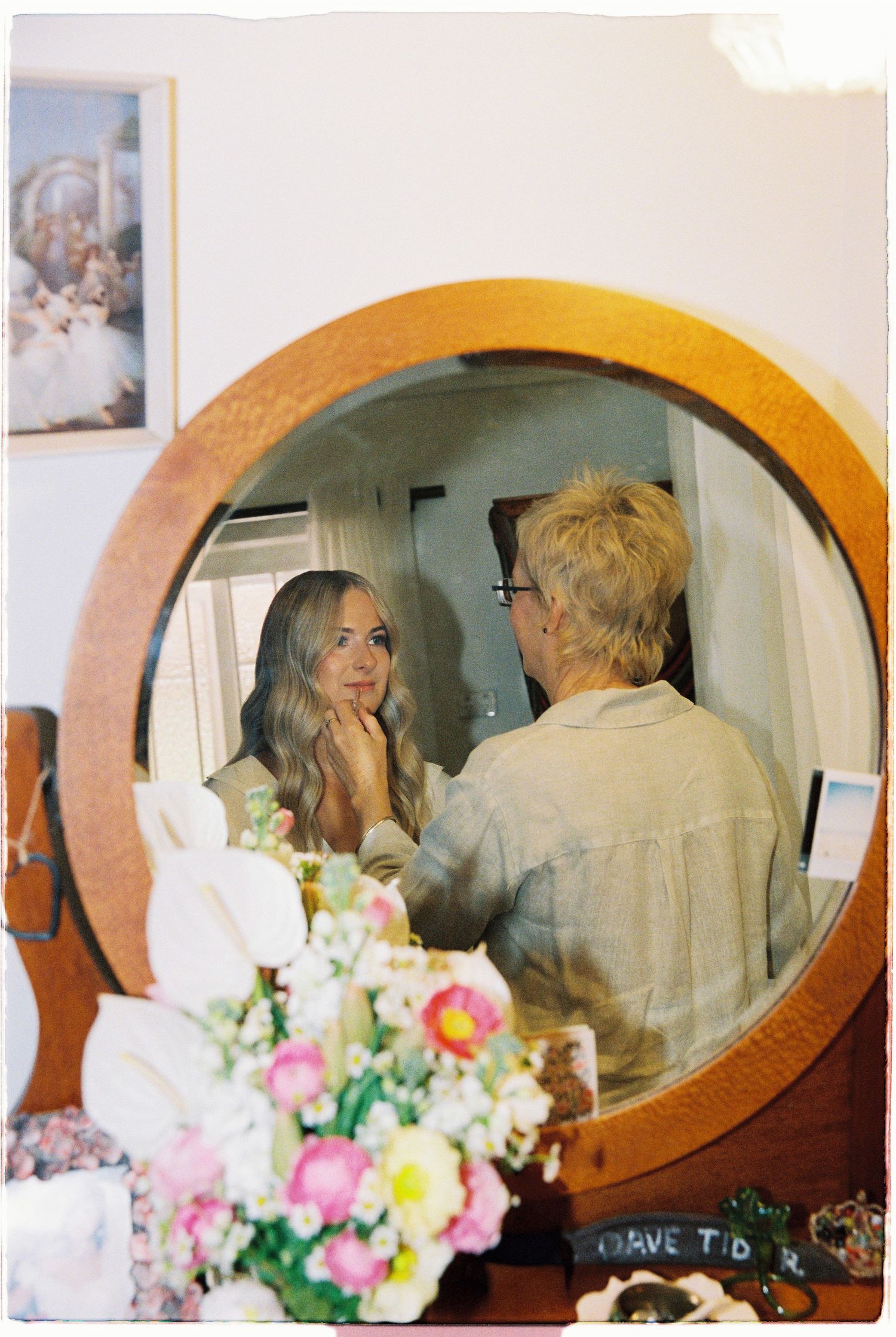 Blonde bride getting makeup done reflected in round wooden mirror with flowers in foreground.