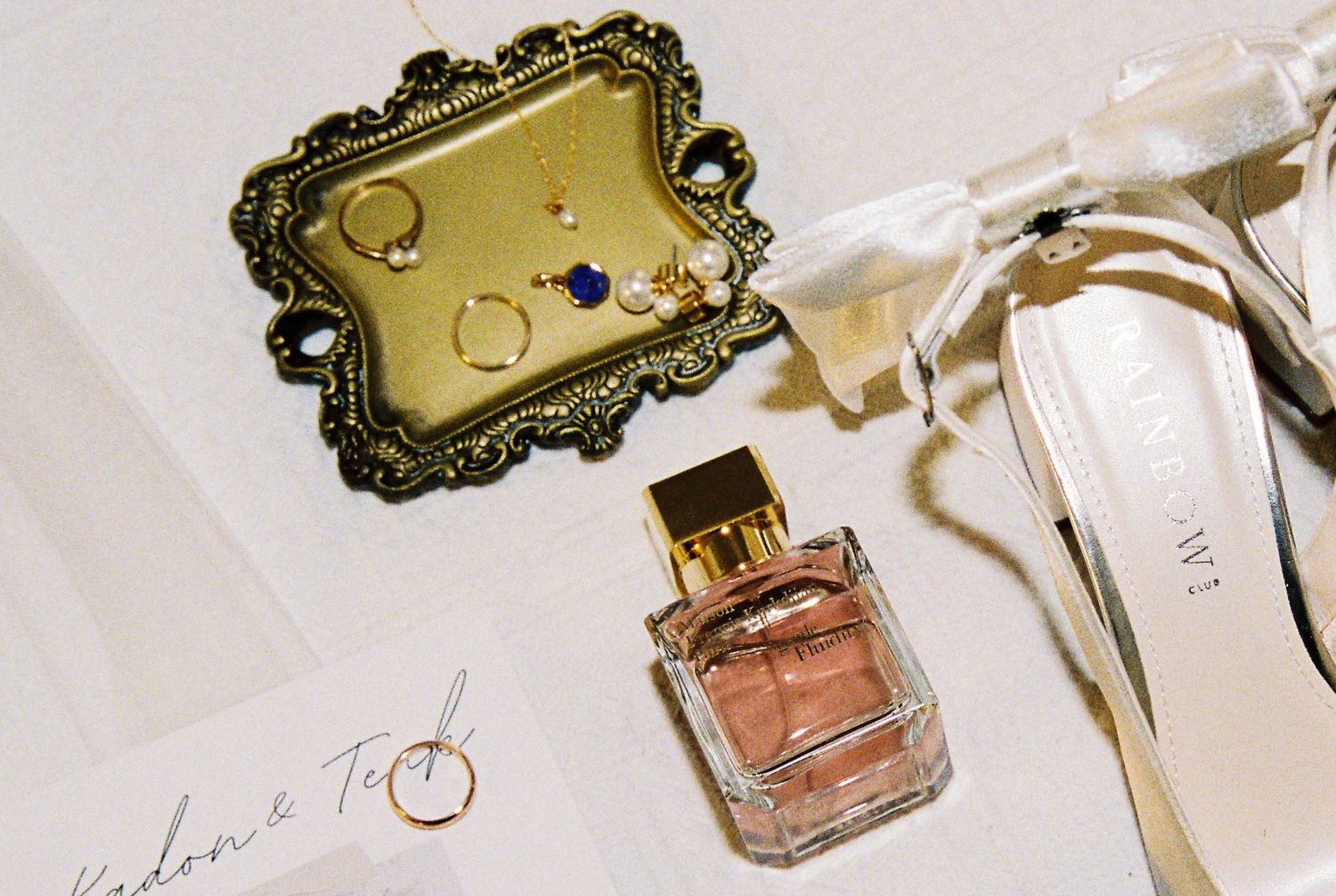Wedding flat lay with perfume bottle, gold jewelry tray, rings, and bridal heels on white surface.