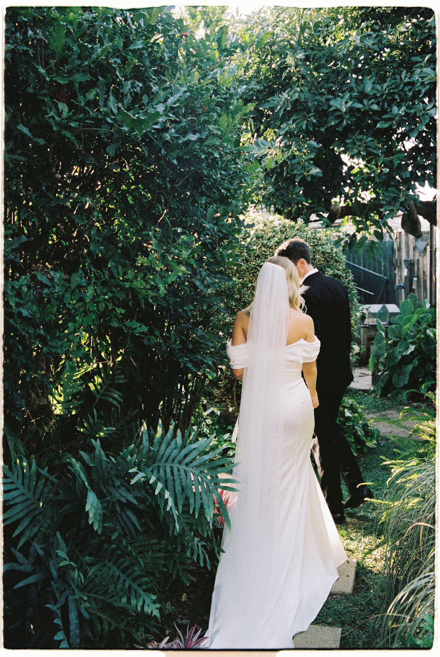Bride in elegant white off-shoulder gown and veil walks with groom through lush green garden pathway.