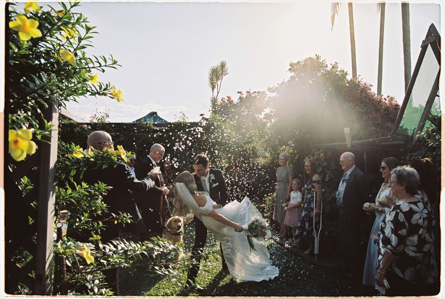 Newlyweds celebrate with confetti shower surrounded by joyful guests in sunlit garden after ceremony.