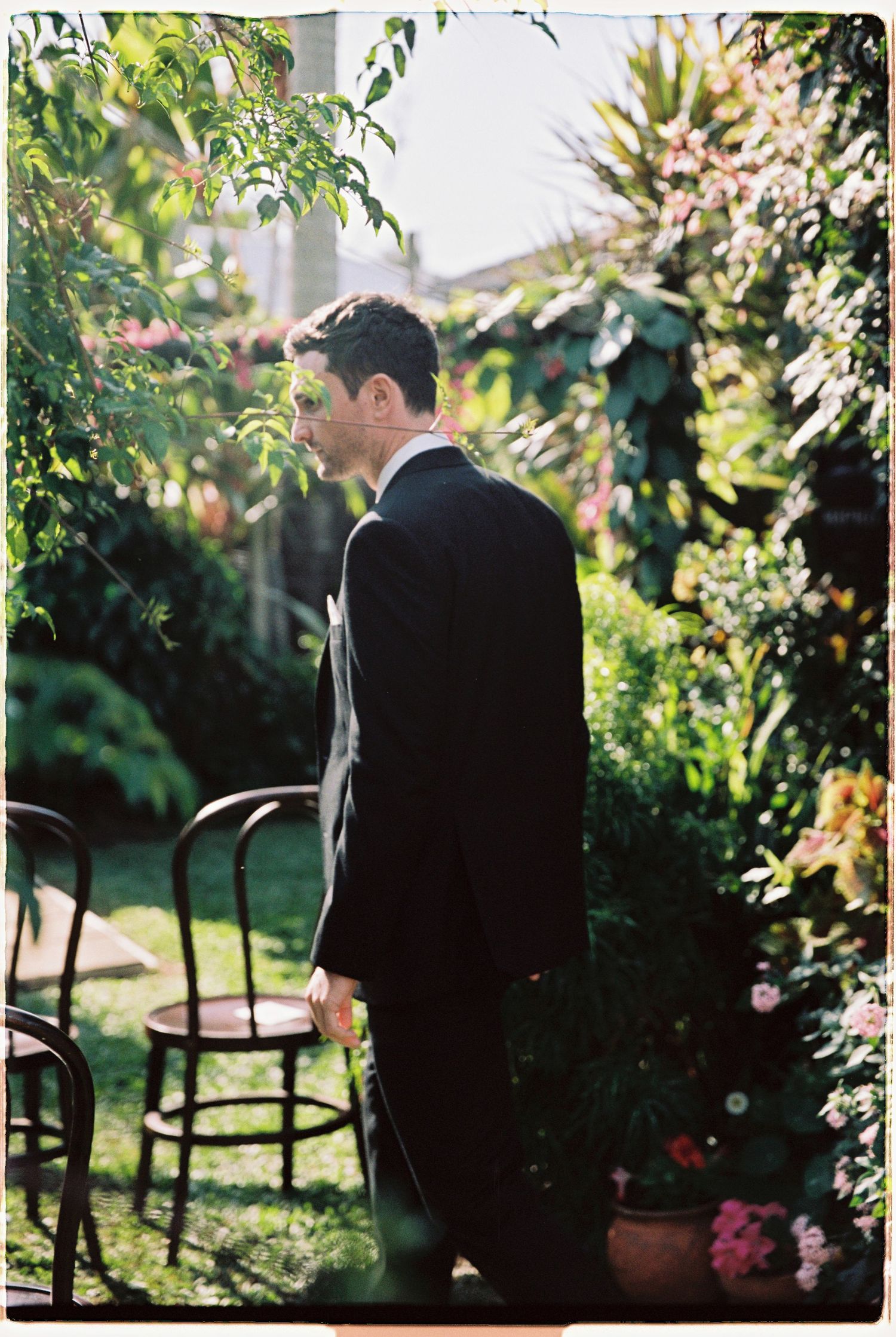 Groom in black suit standing in lush garden ceremony space with wooden chairs and colorful flowers.