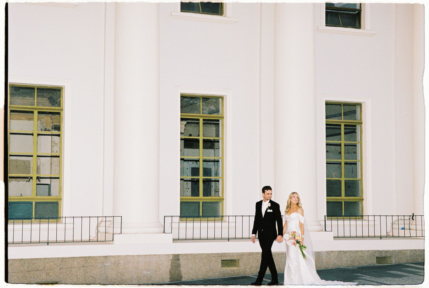 Bride and groom walk together along terrace of grand white classical building with green windows.