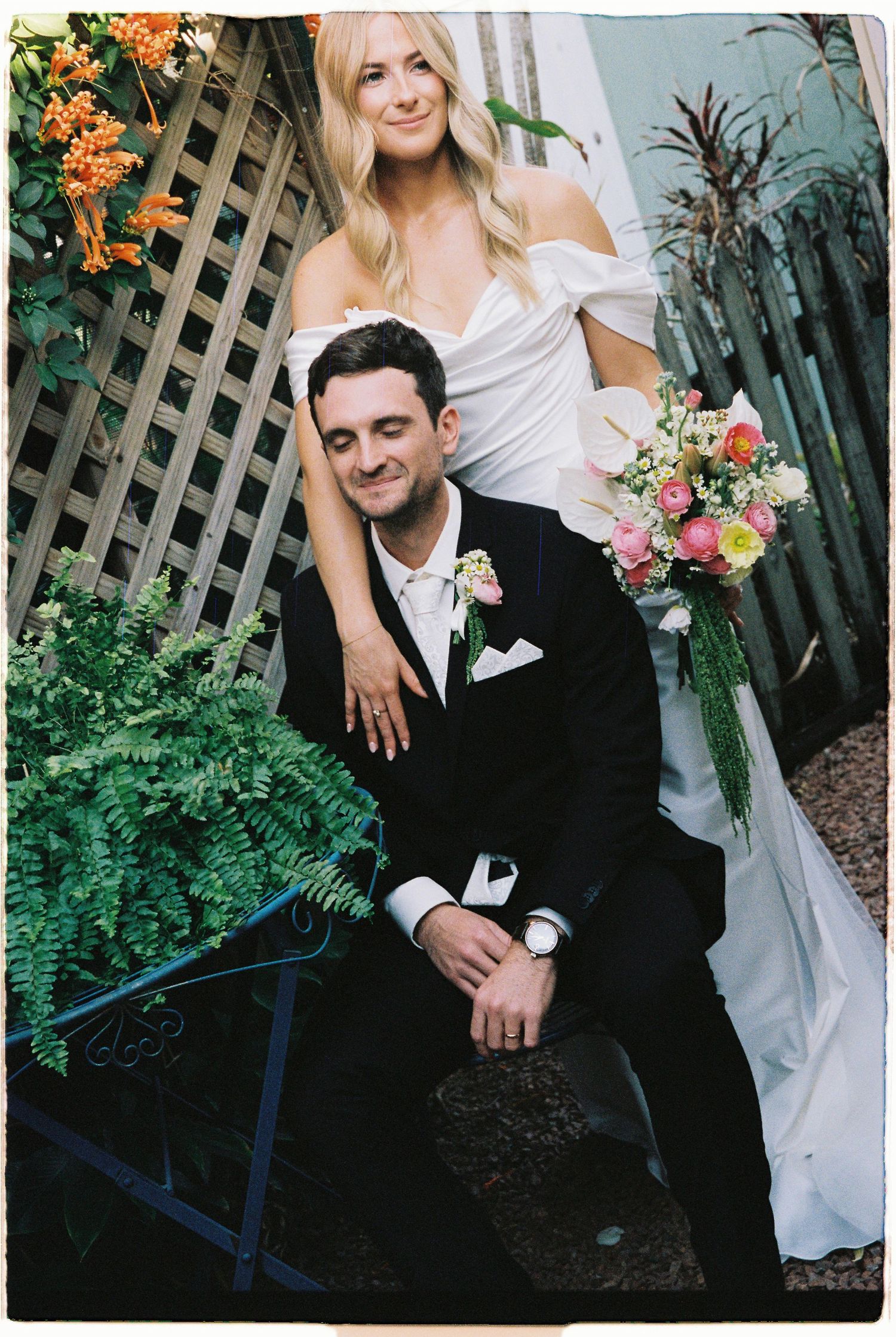 Smiling bride in white off-shoulder gown holds colorful bouquet while posing with seated groom in black tuxedo.