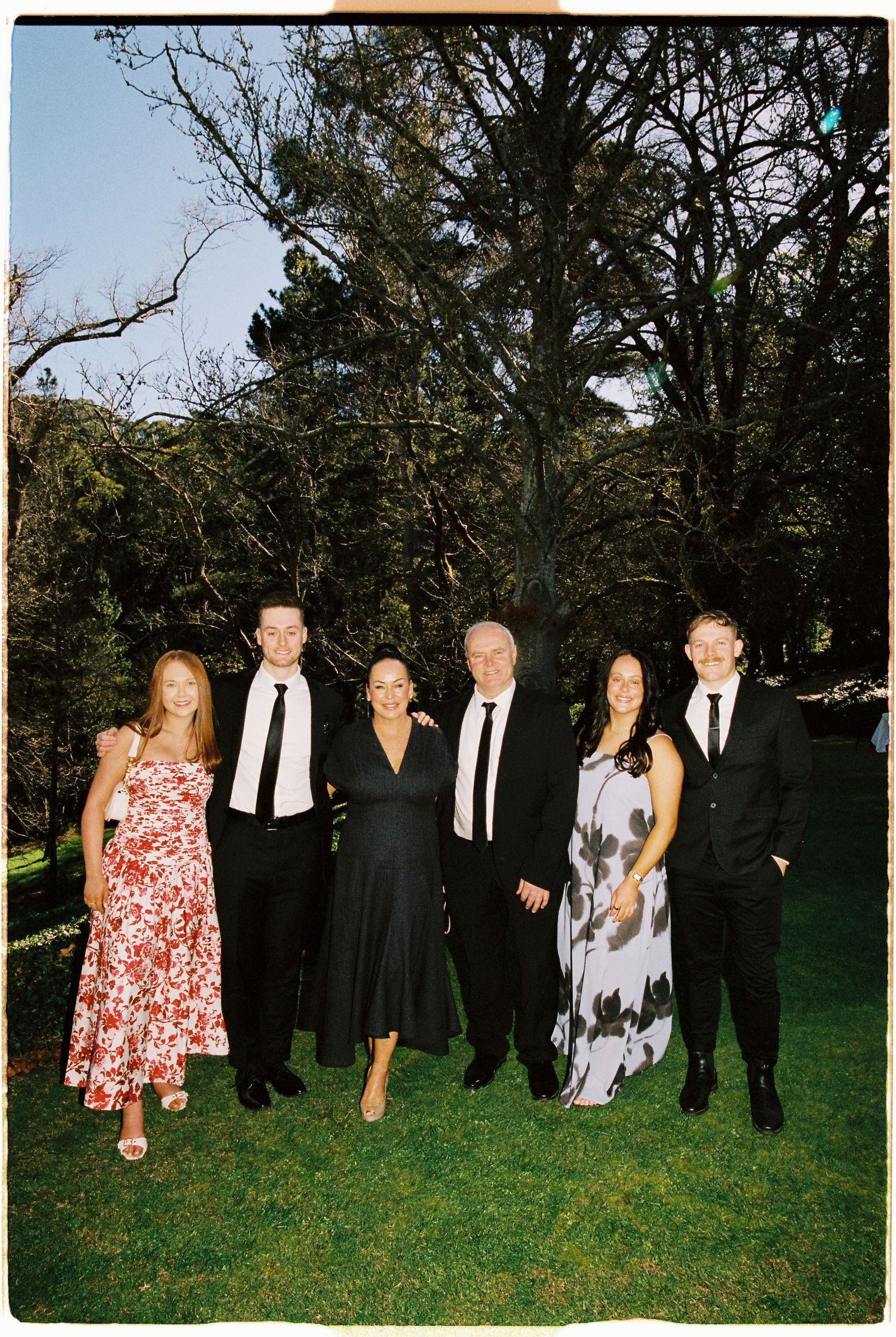 Group of six elegantly dressed wedding guests pose together on a lawn with tall trees in the background.