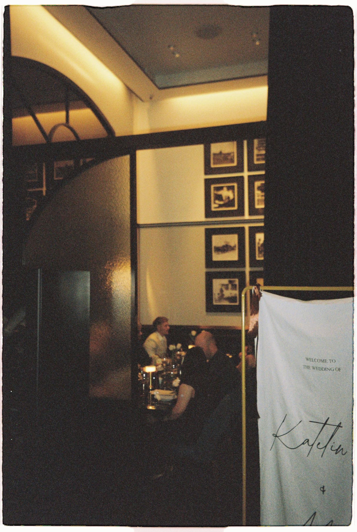 Dimly lit upscale restaurant interior with diners seated at tables, framed artwork on walls, and a welcome sign in the foreground.